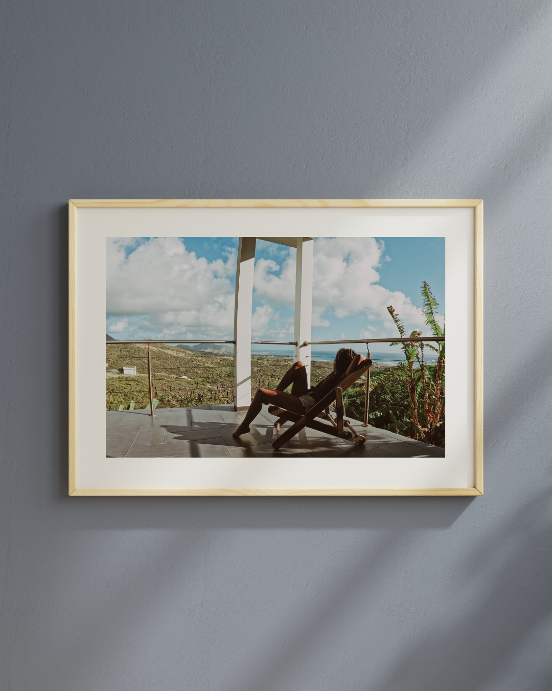 Photo of a woman relaxing on a lounge chair on a balcony overlooking a green hill and sea, under a partly cloudy sky.