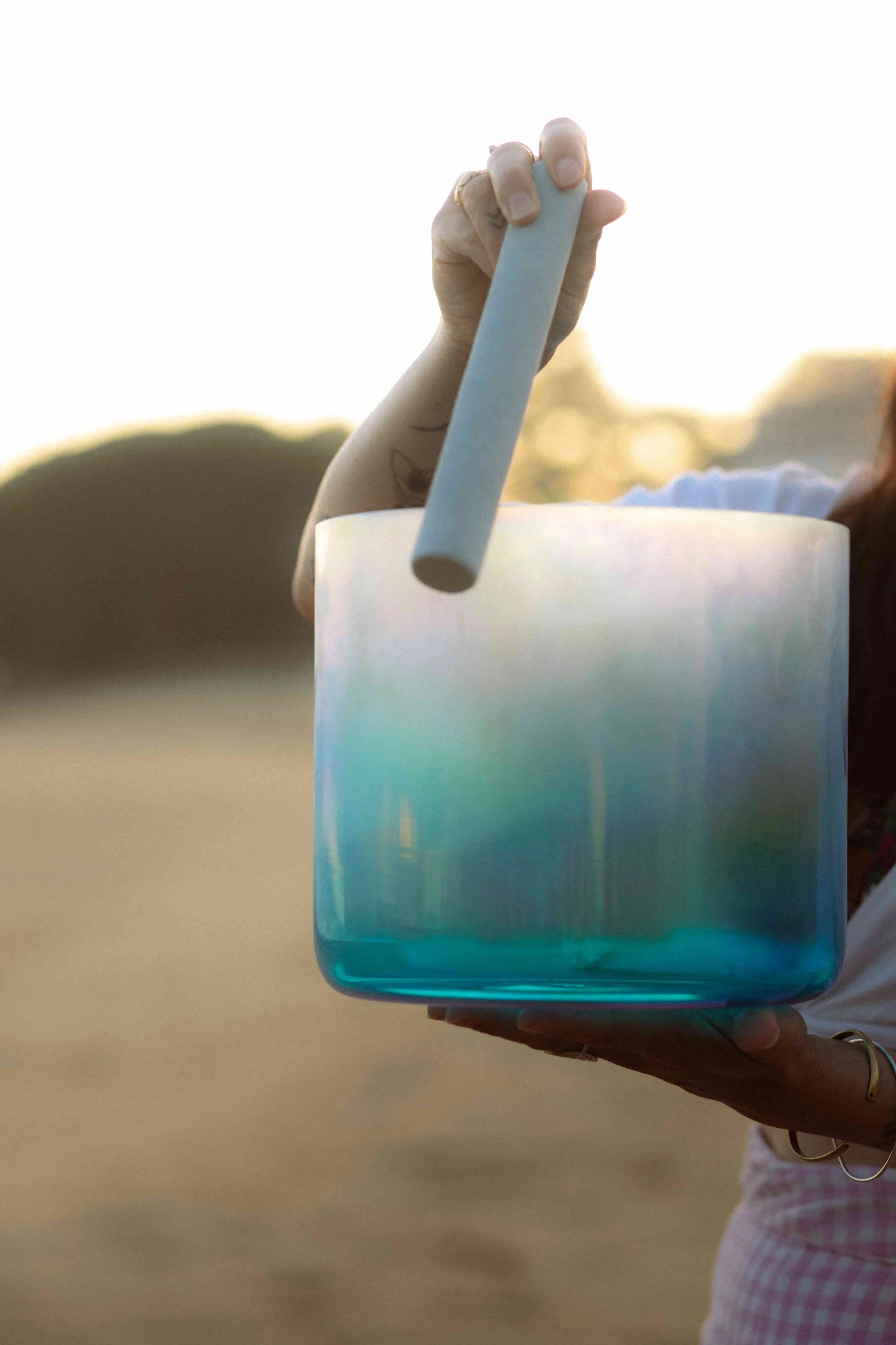Laura, praticienne en bains sonores, présentant un bol alchimique au bord de la mer au coucher du soleil, rituel de son et d’énergie.
