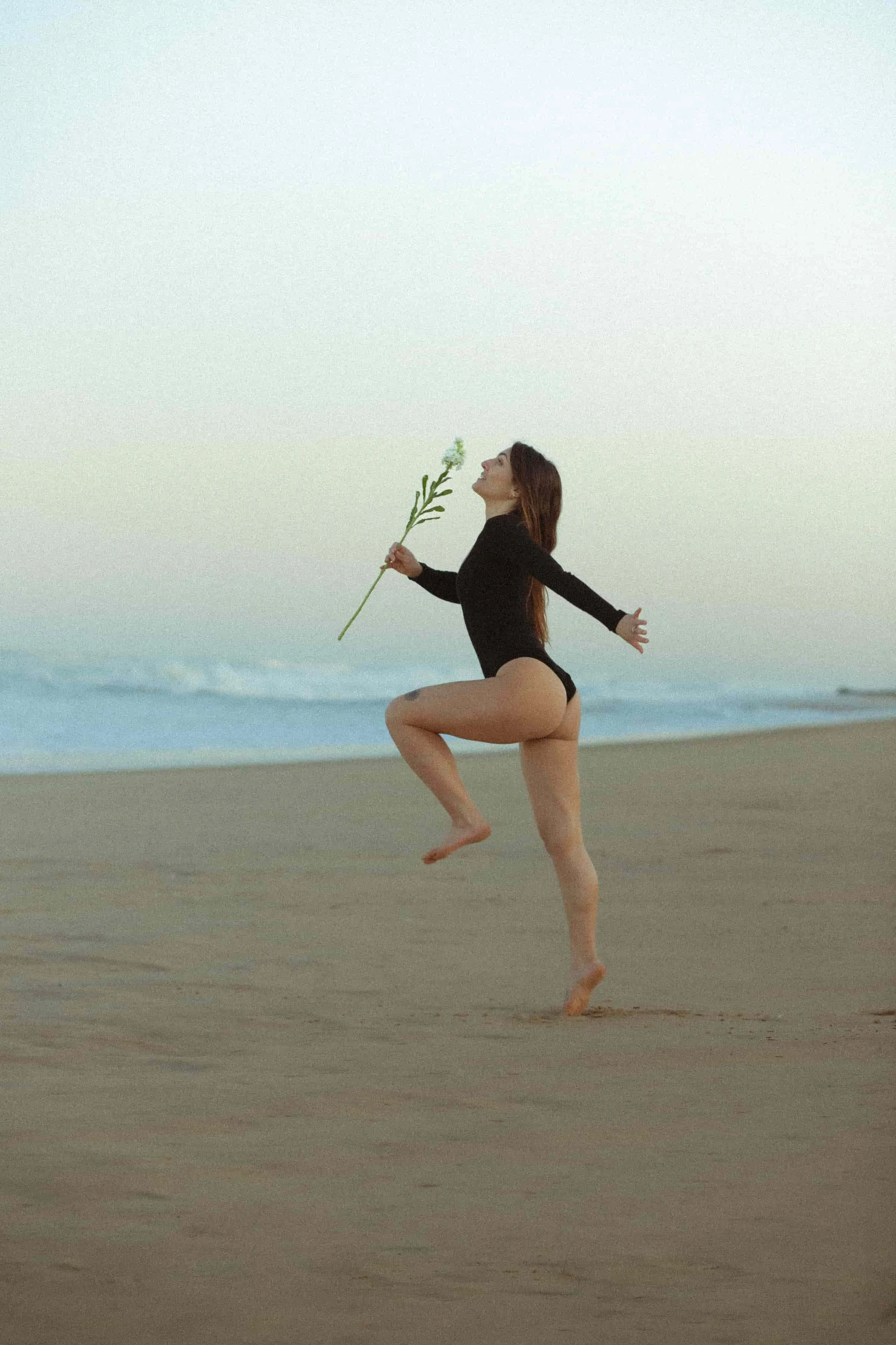 Photo artistique en noir et blanc d’une femme sautant sur la plage, tenant une fleur à la main, symbole de liberté et de connexion à l’océan.