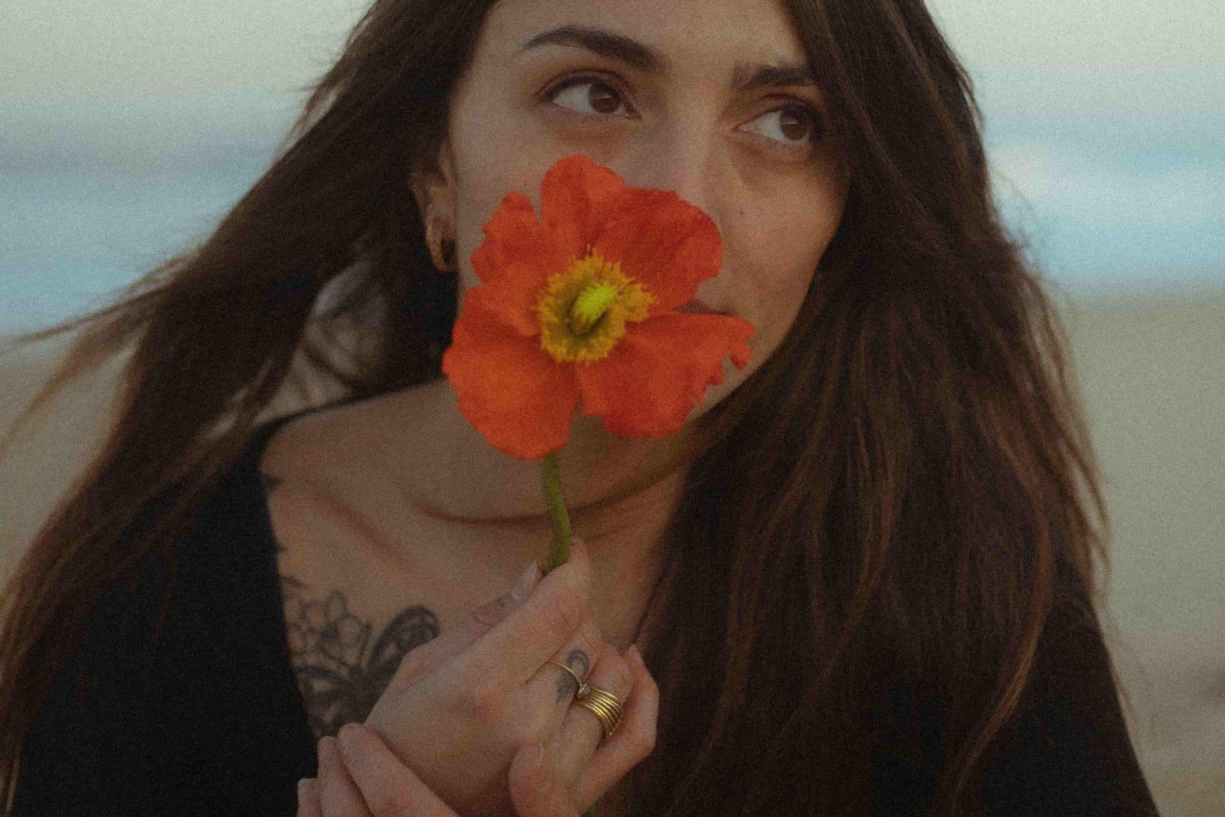 Laura, fleuriste freelance, tenant une fleur orange face à l’océan, expression de son lien créatif avec la nature.