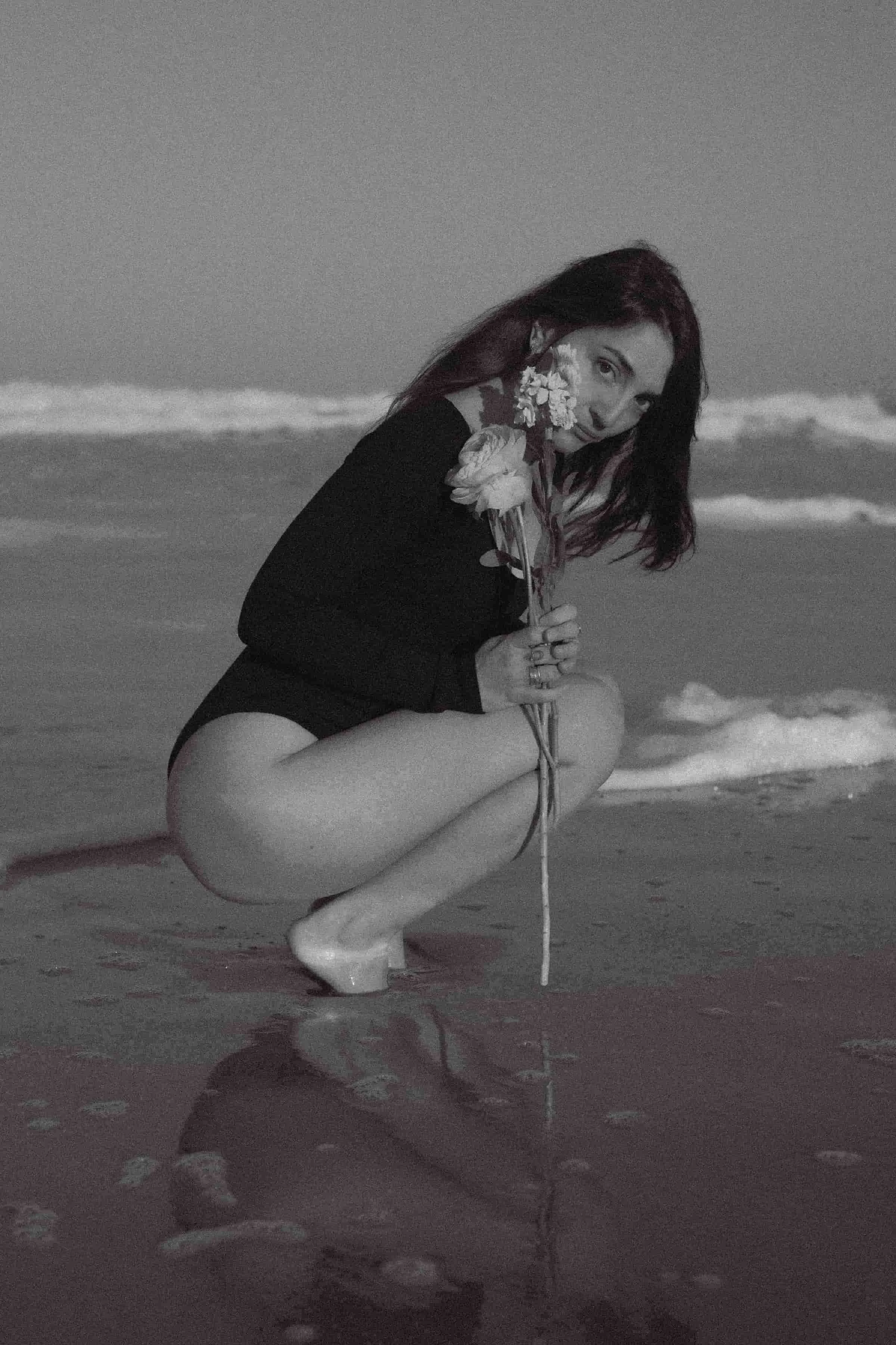 Laura, créatrice multidisciplinaire : bains sonores, conception web et art floral, tenant un bouquet de fleurs face à la mer, photo en noir et blanc.