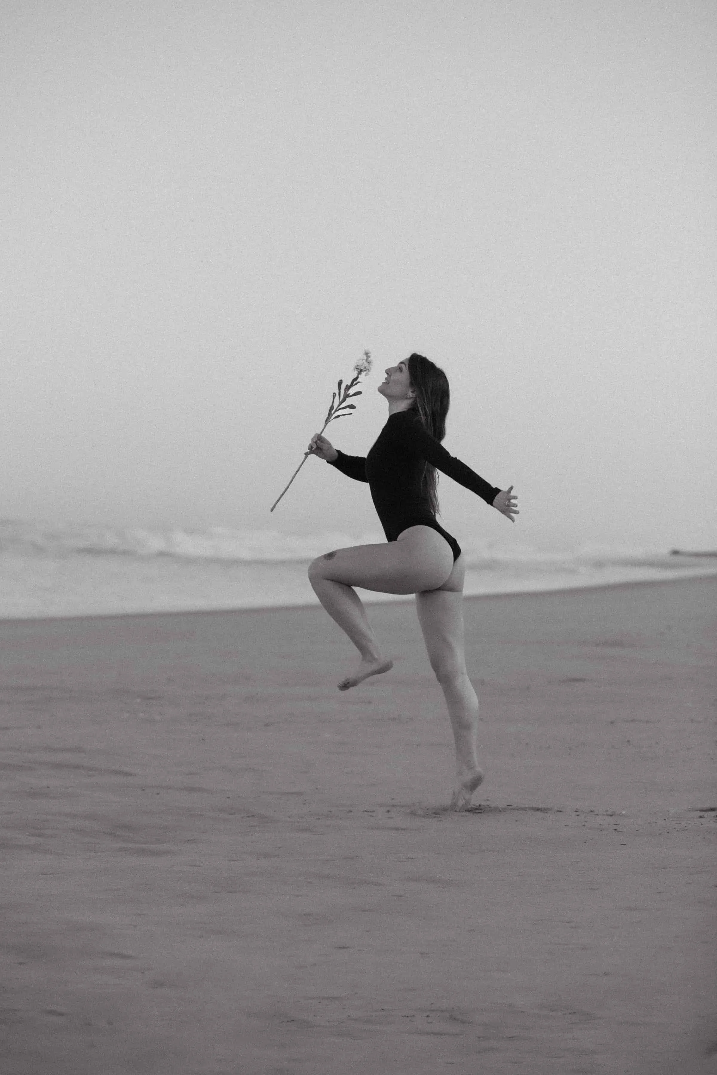 Photo artistique en noir et blanc d’une femme sautant sur la plage, tenant une fleur à la main, symbole de liberté et de connexion à l’océan.