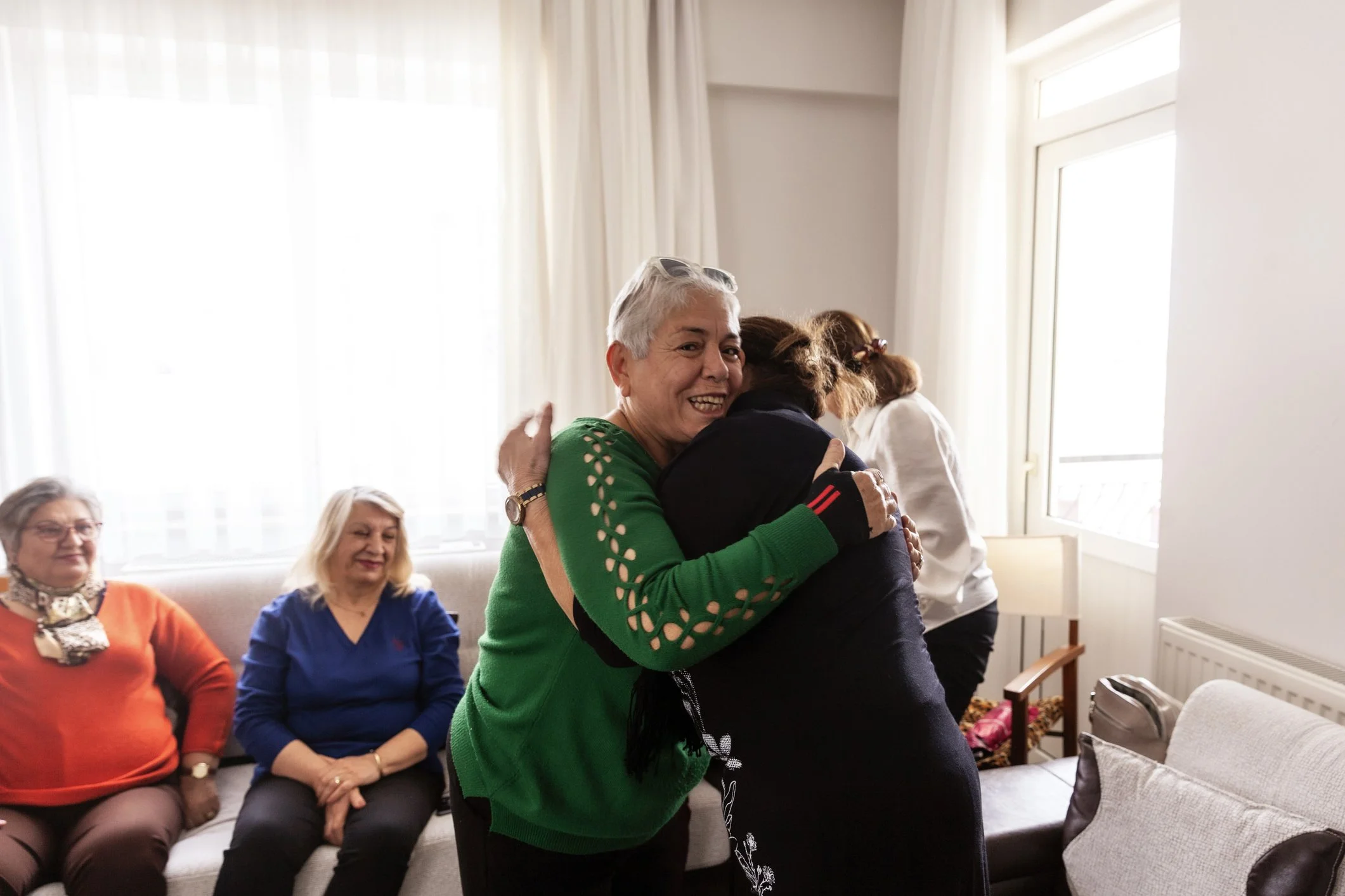 Two women hugging in a brightly lit room with several seated women watching. The women hugging have gray and brown hair, and are smiling. The room has large windows with white curtains and a comfortable sitting area.