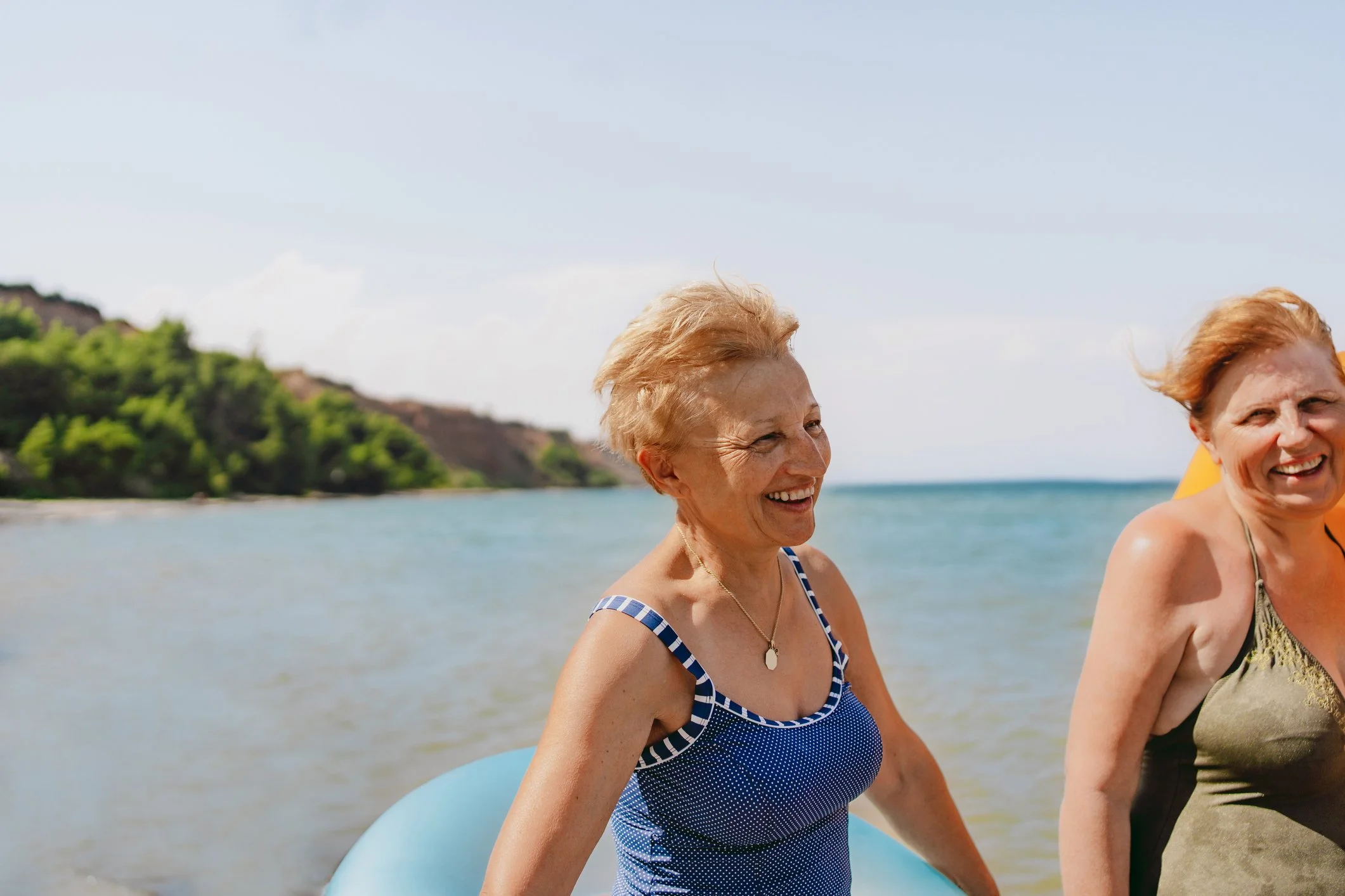 Two women smiling and walking on the beach near the ocean, with green hills in the background, under a blue sky.