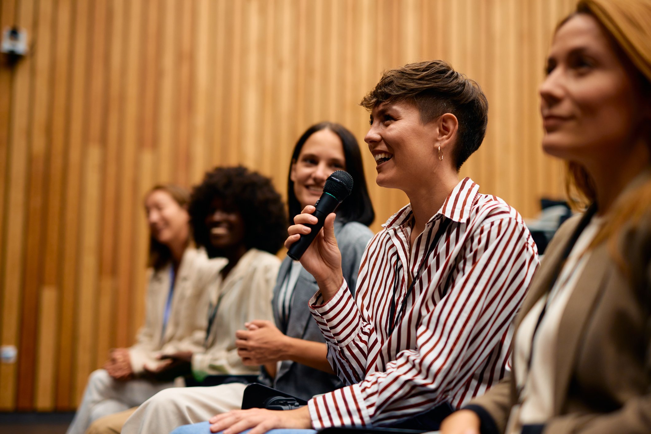 A woman smiling and holding a microphone in a panel discussion, sitting among other women in an auditorium with wooden paneling in the background.