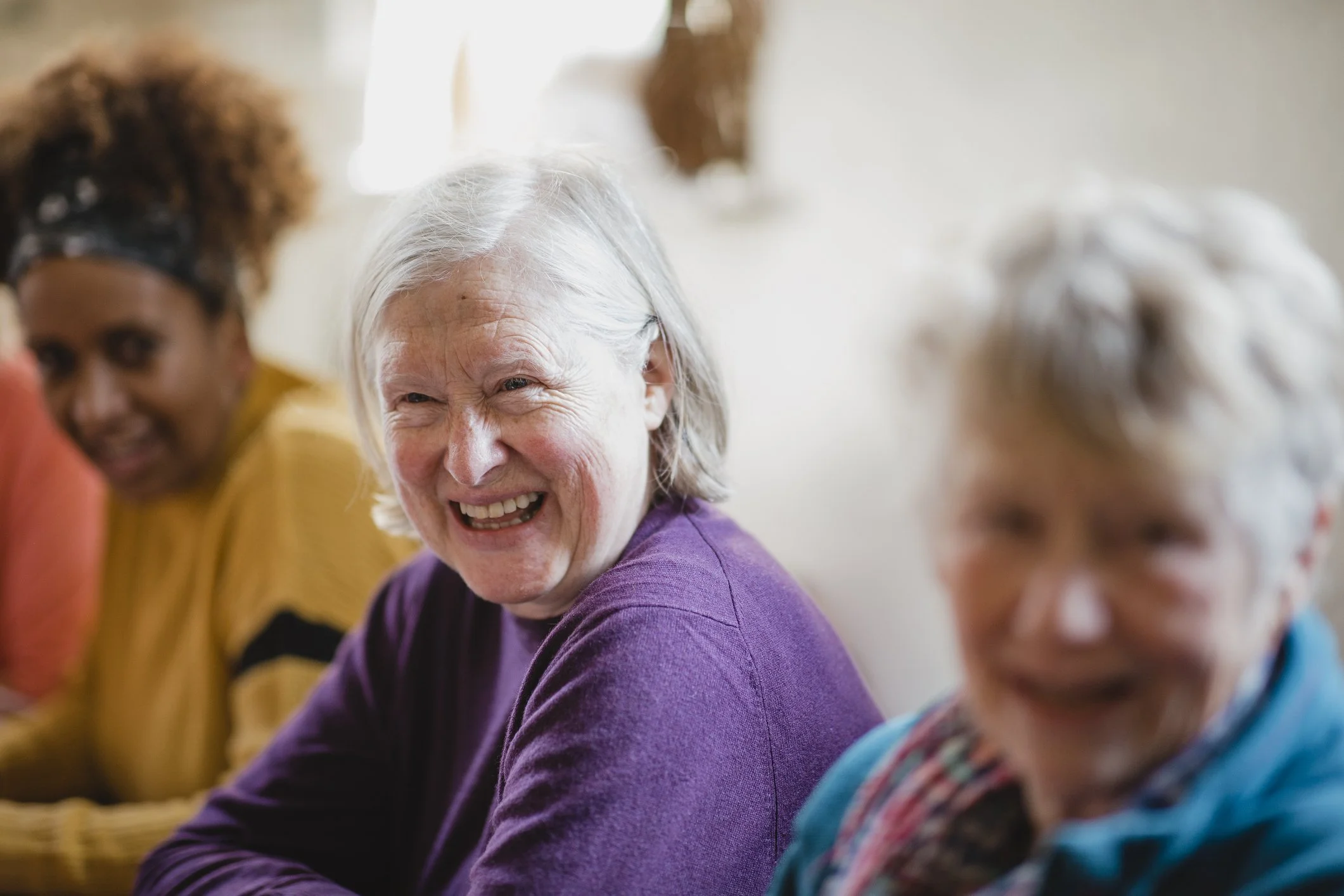A group of elderly women sitting together, smiling and talking in a bright indoor space.