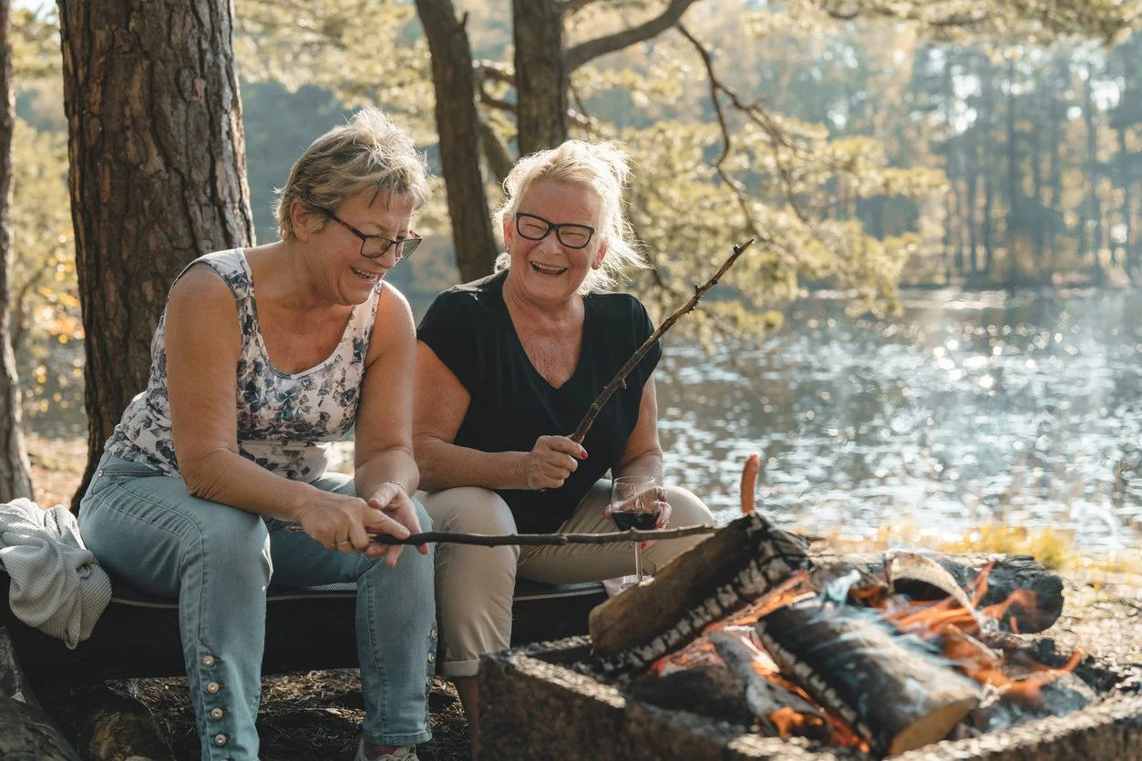 Two women sitting by a campfire near a lake, enjoying drinks and laughs.