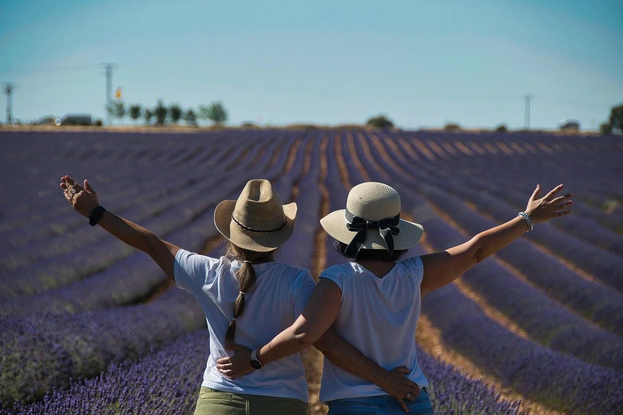 Two women wearing sun hats and white shirts standing in a lavender field, arms outstretched, enjoying the view.