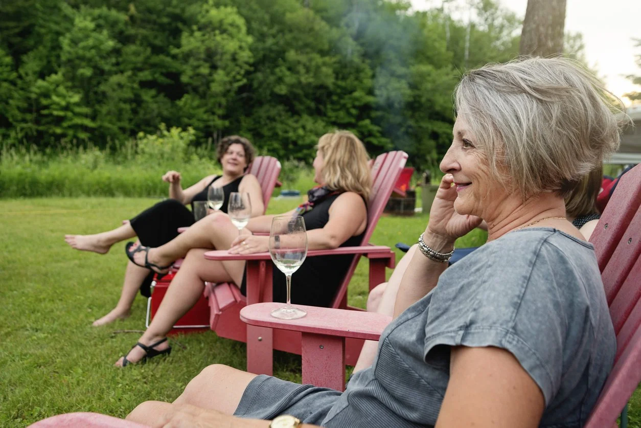 Four women relaxing outdoors on pink Adirondack chairs around sunset, with glasses of wine on a grassy area near trees.