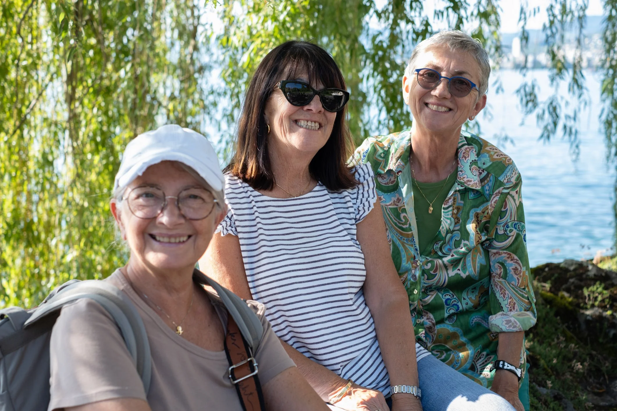 Four women sitting outdoors near a body of water, smiling, with trees in the background. One woman in the front is wearing glasses and a white hat, another in the middle has dark hair and sunglasses, and the two others have short hair and sunglasses.