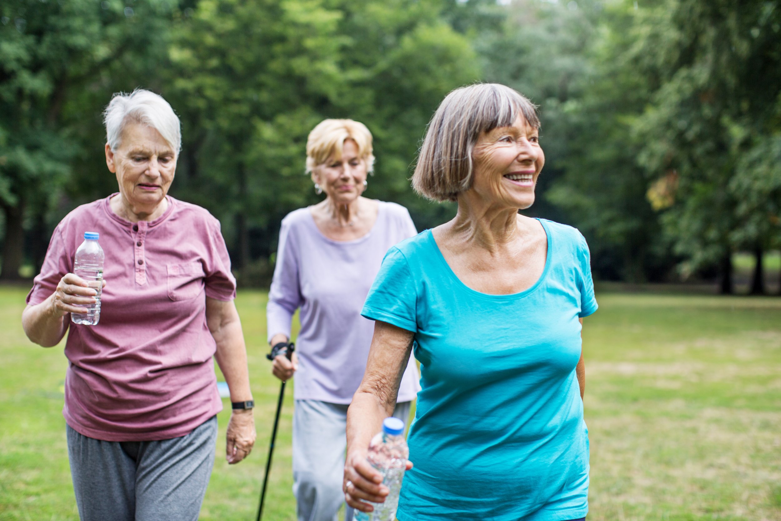 Three elderly women walking outdoors in a park, holding water bottles, with trees in the background.