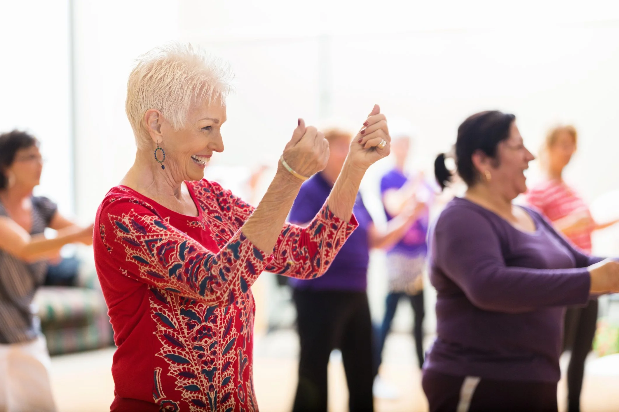 An elderly woman with short white hair, wearing a red patterned top and earrings, is dancing or exercising with others in a bright room.