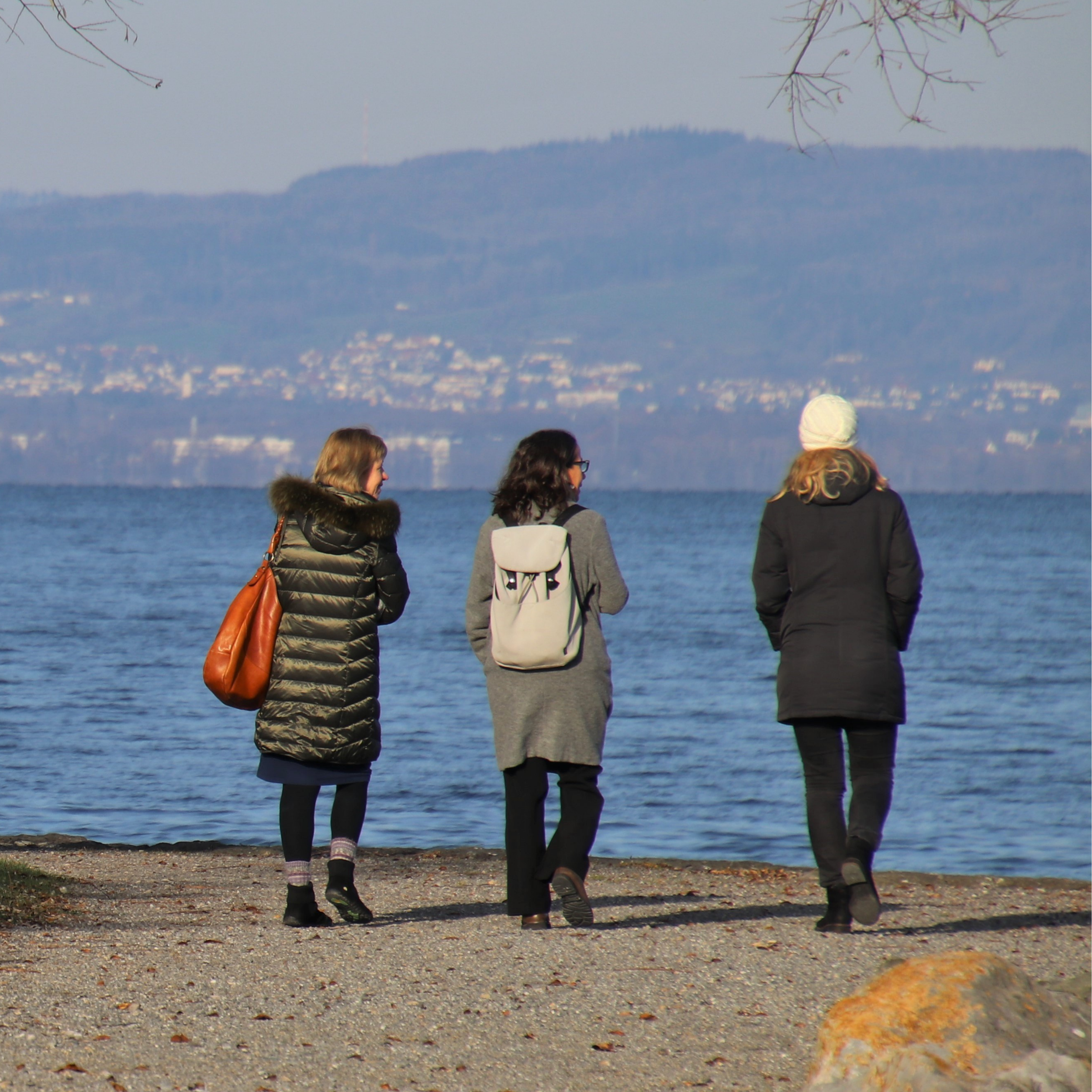 Four women walk along a beach with water and distant hills in the background. They are dressed in warm clothing, and some are carrying bags.