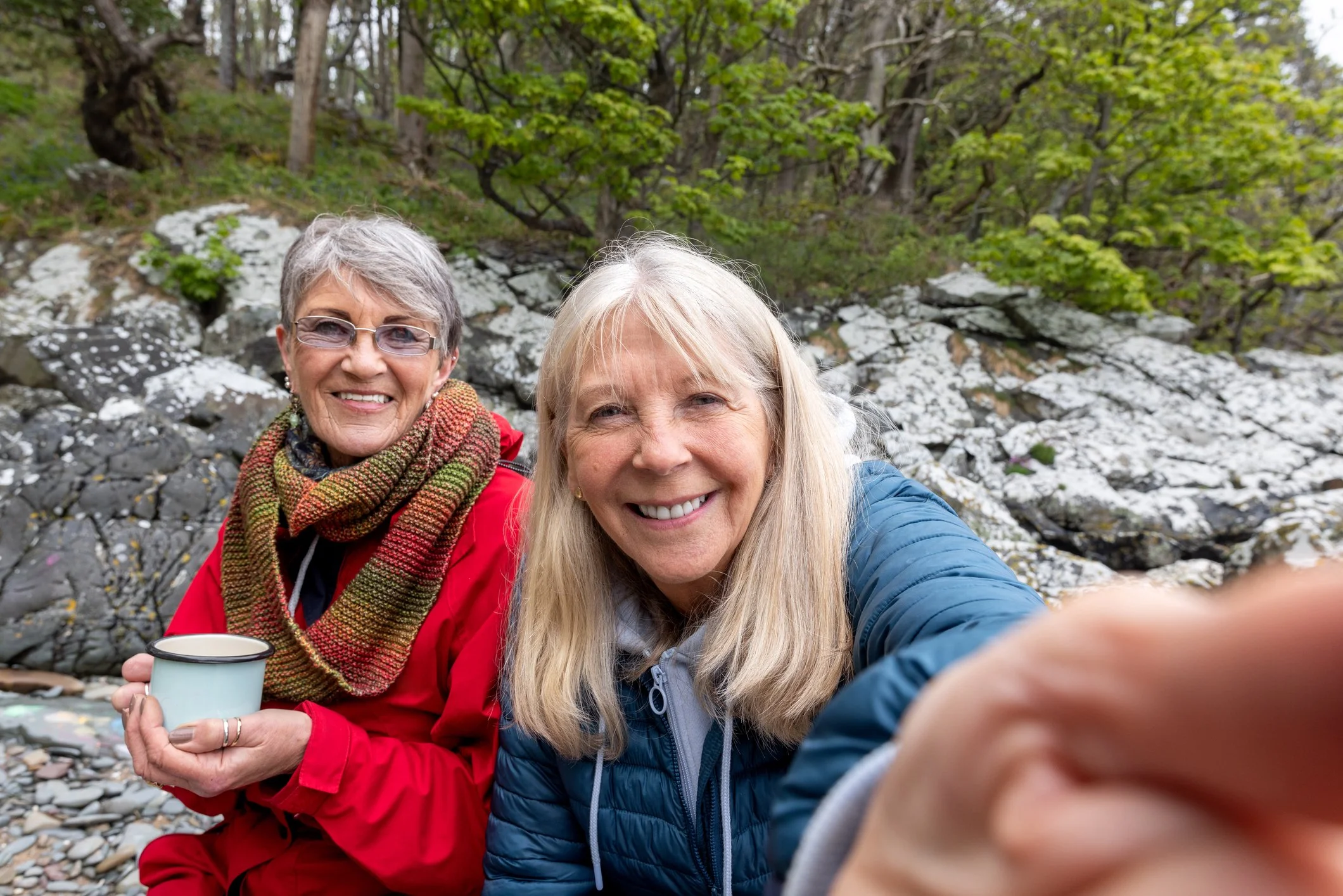Two smiling women taking a selfie outdoors near a rocky stream with trees and green foliage in the background.