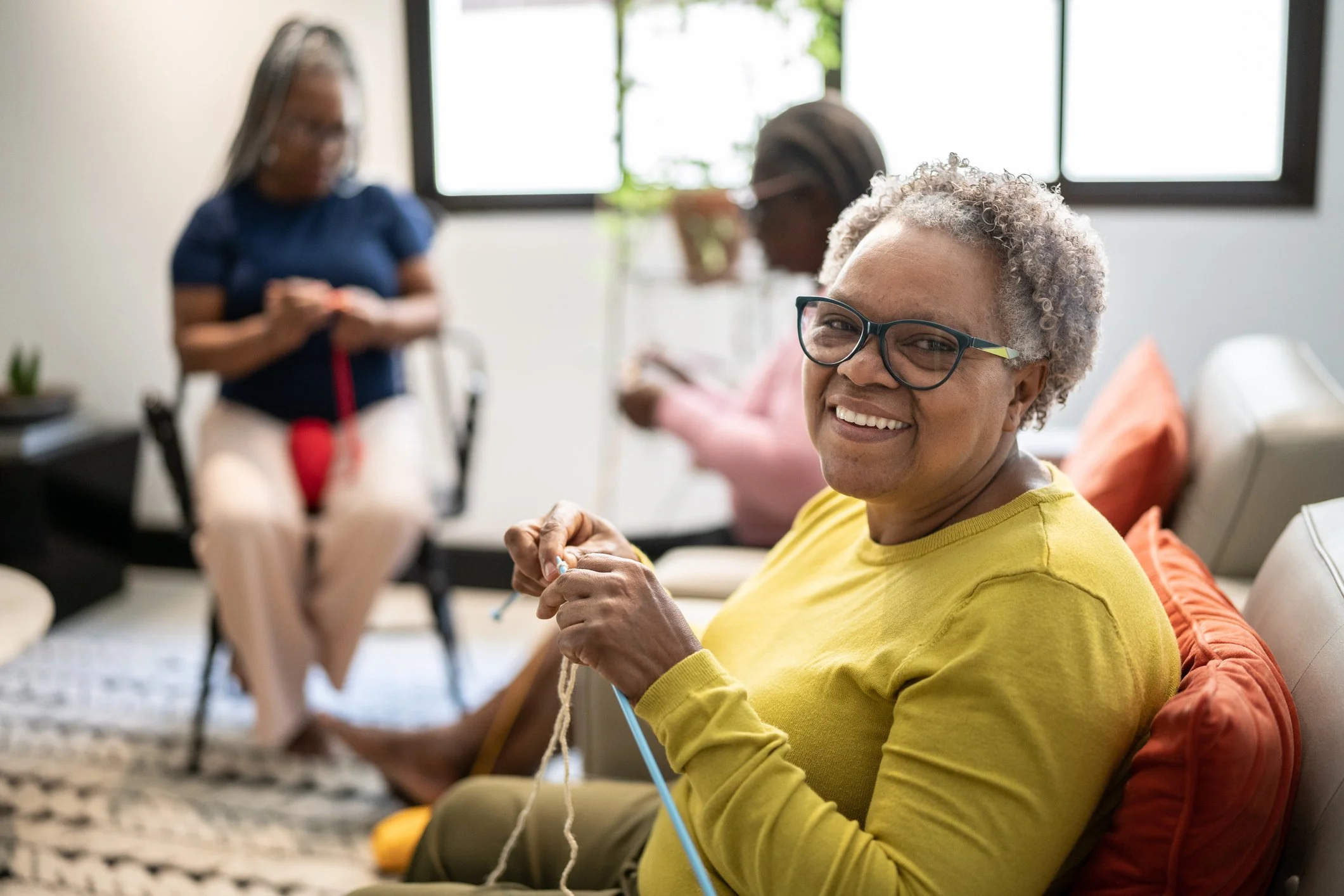 An elderly woman with gray curly hair and glasses smiling while knitting in a bright, modern living room with large windows, orange cushions, and a patterned rug, with two blurred women in the background.