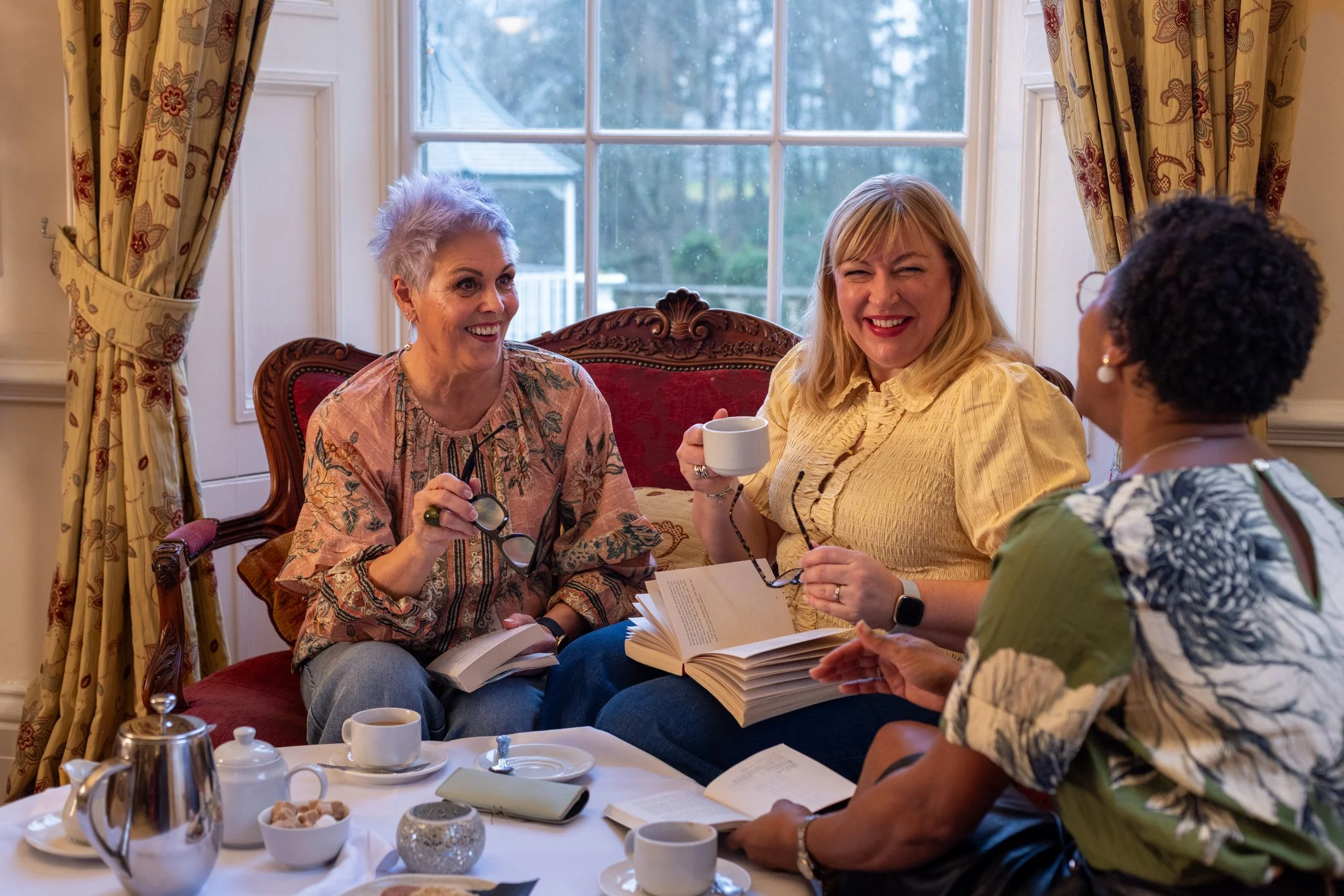 Four women sitting at a dining table during a gathering, laughing and engaging in conversation, with cups, a teapot, and snacks on the table, inside a room with a large window and colorful curtains.