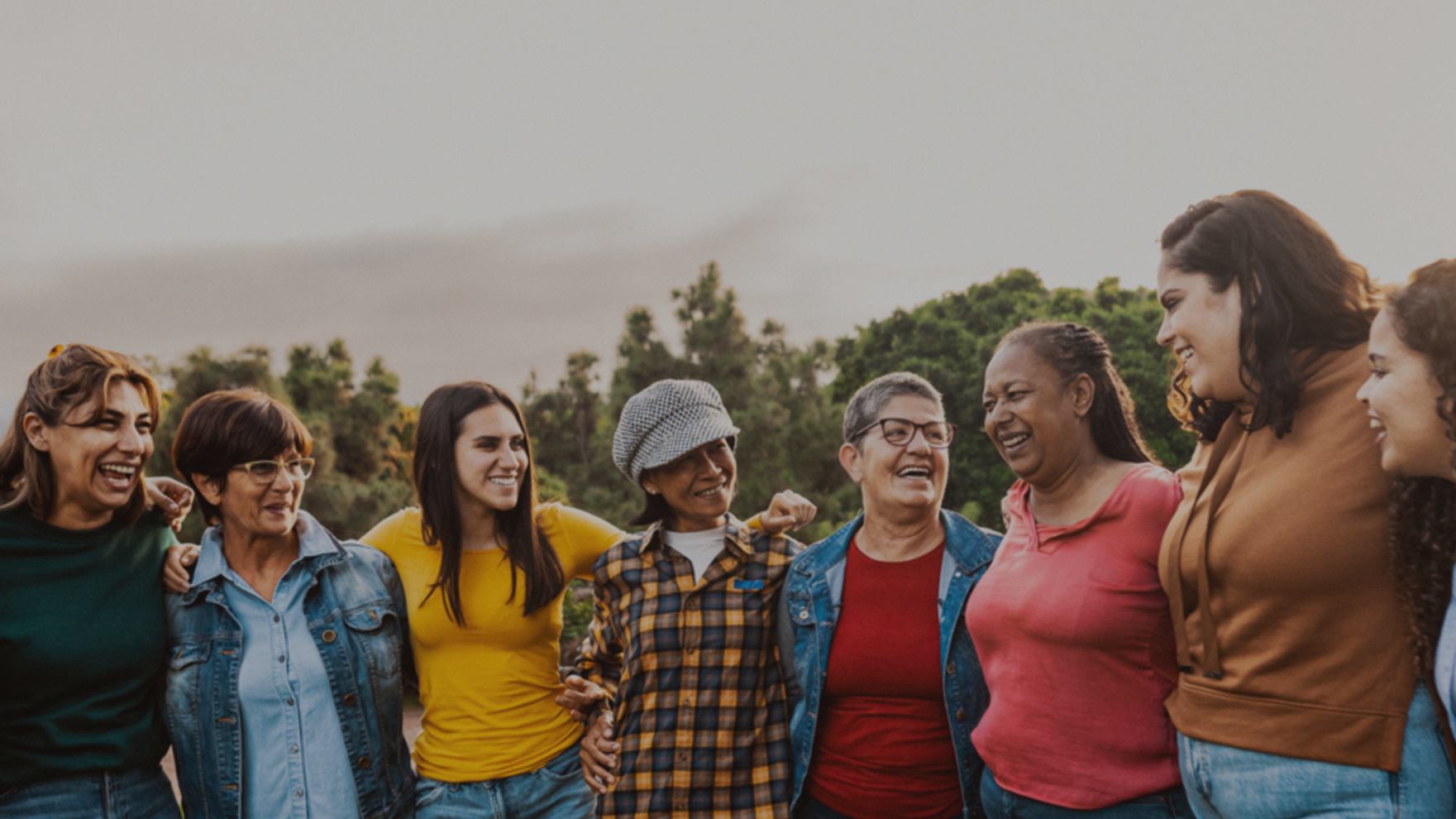 Group of women standing outdoors, smiling and embracing each other, with trees in the background.