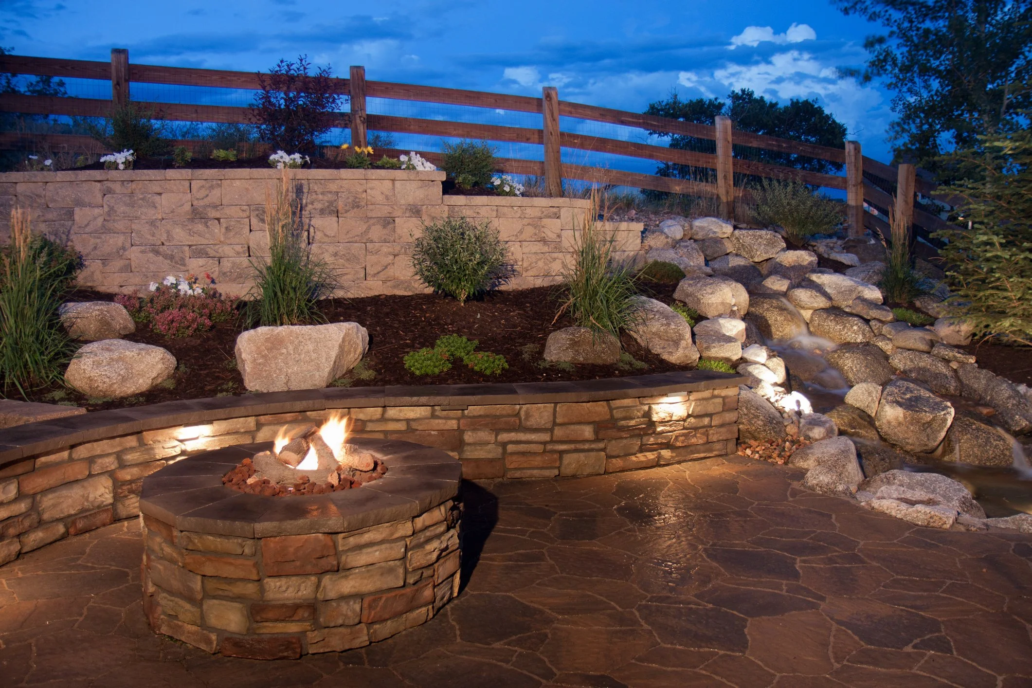 Backyard with a fire pit, stone retaining wall, plants, rocks, and a wooden fence under a cloudy evening sky.