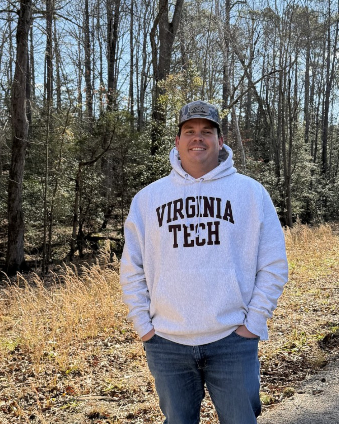 A man standing outdoors in a forested area during fall, wearing a white hoodie with 'VIRGINIA TECH' written on it, a camouflage cap, and jeans.
