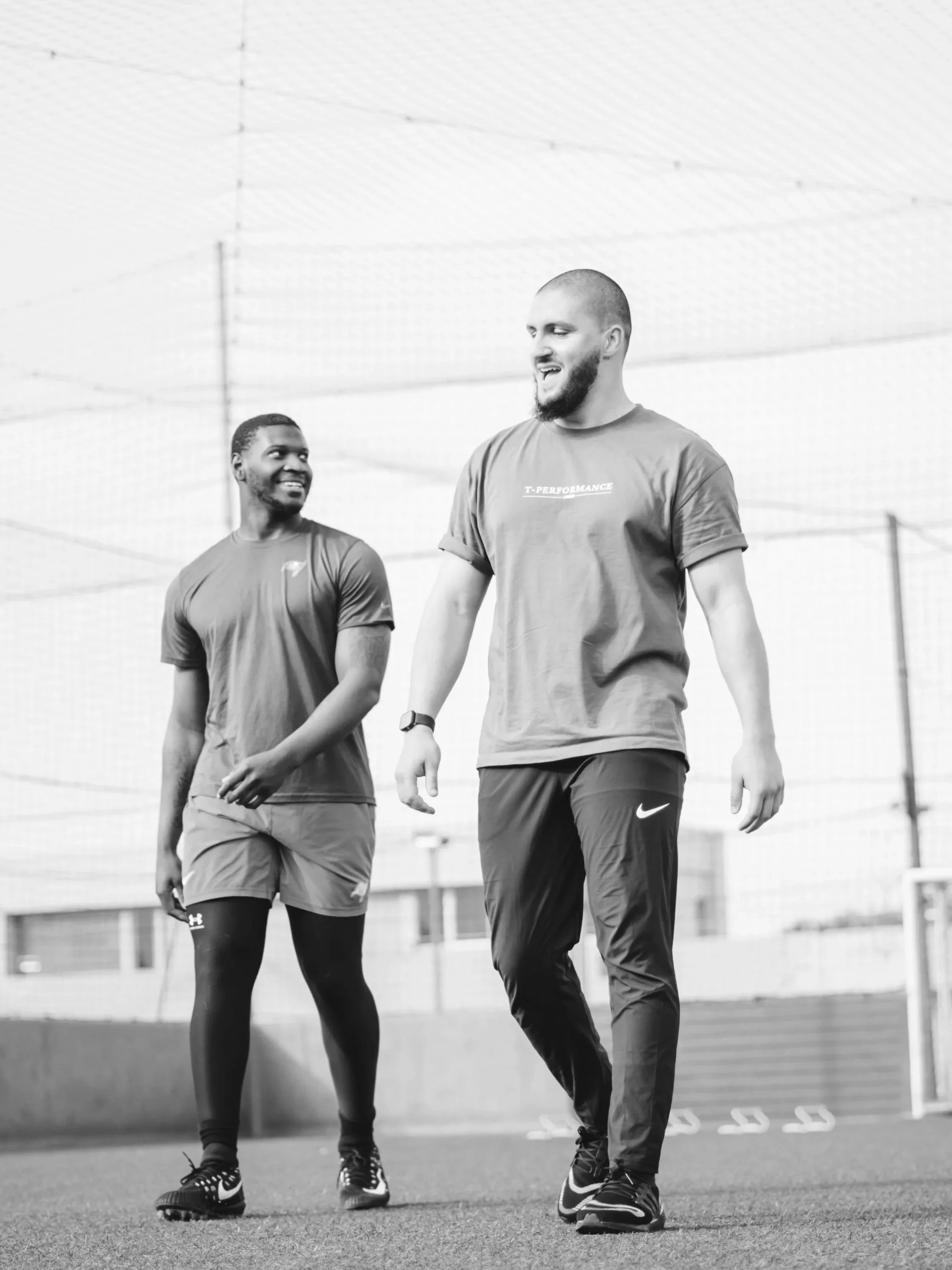 Two men walking and talking on a sports field with a netted structure overhead, both dressed in athletic clothing, smiling and interacting.