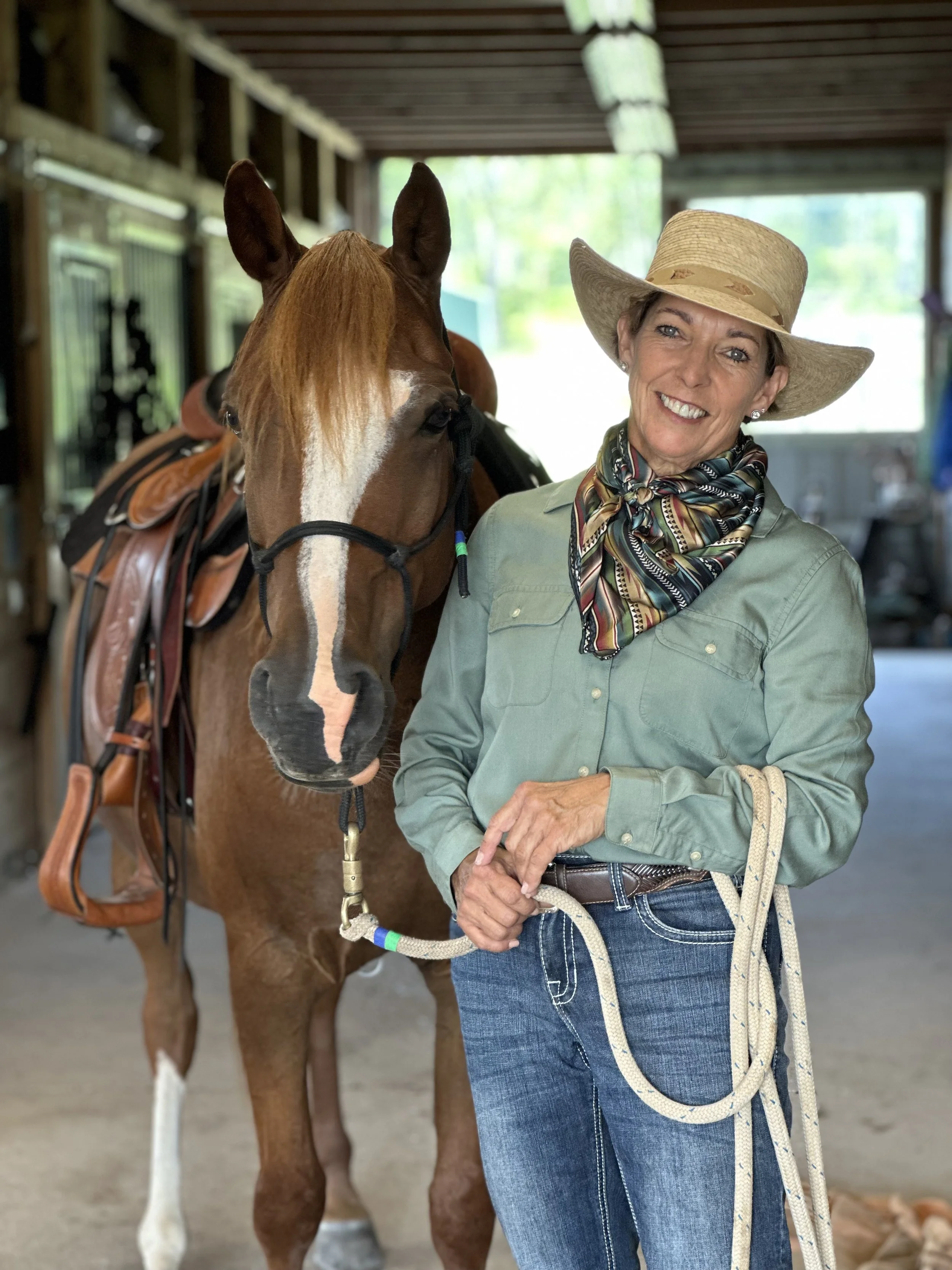 A woman in cowgirl attire, wearing a wide-brimmed cowboy hat and bandana, standing next to a brown horse with a western saddle inside a clean barn ready to show in Cowboy Dressage.