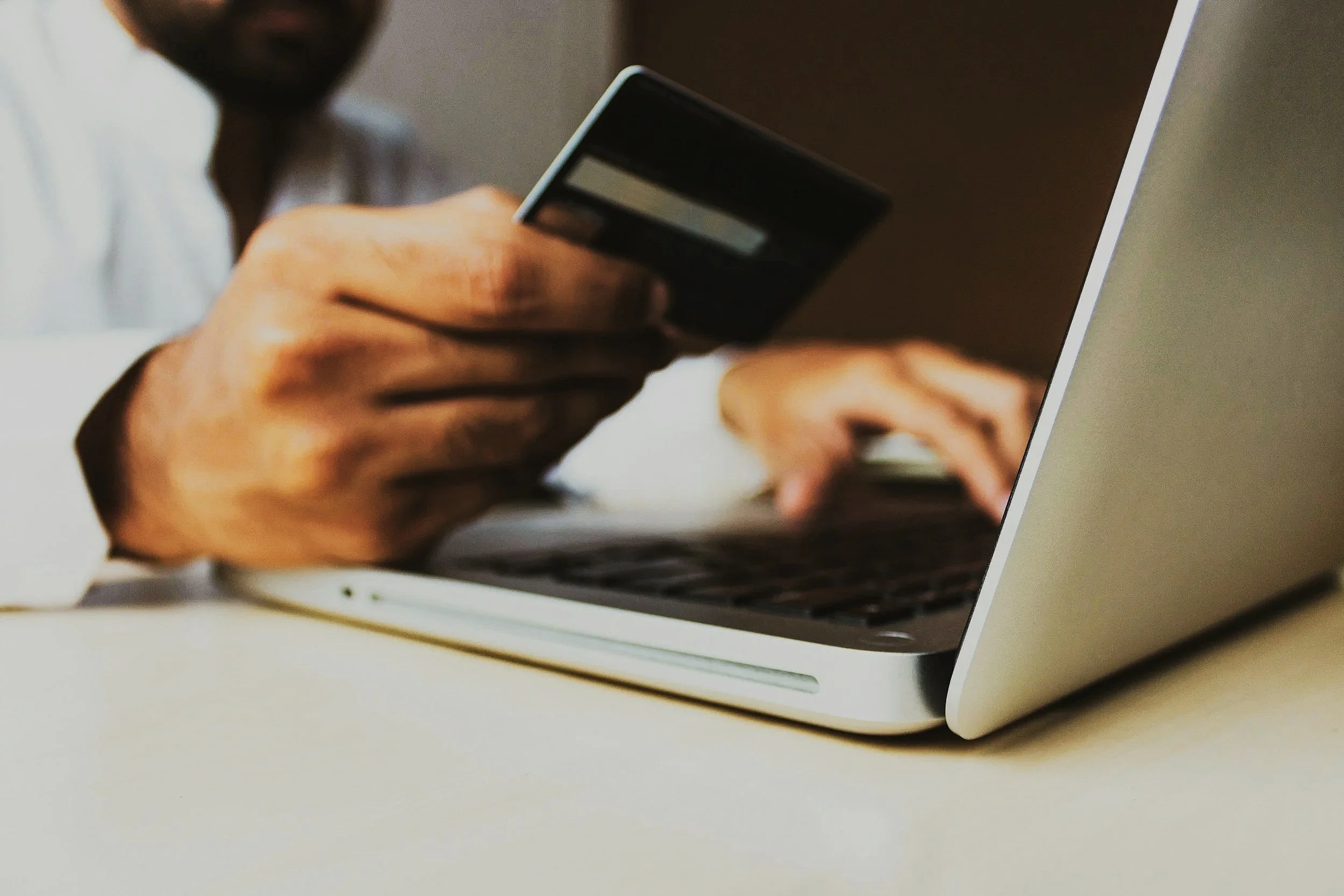 Close-up of a person holding a credit card near a laptop keyboard with a hand on the laptop trackpad.