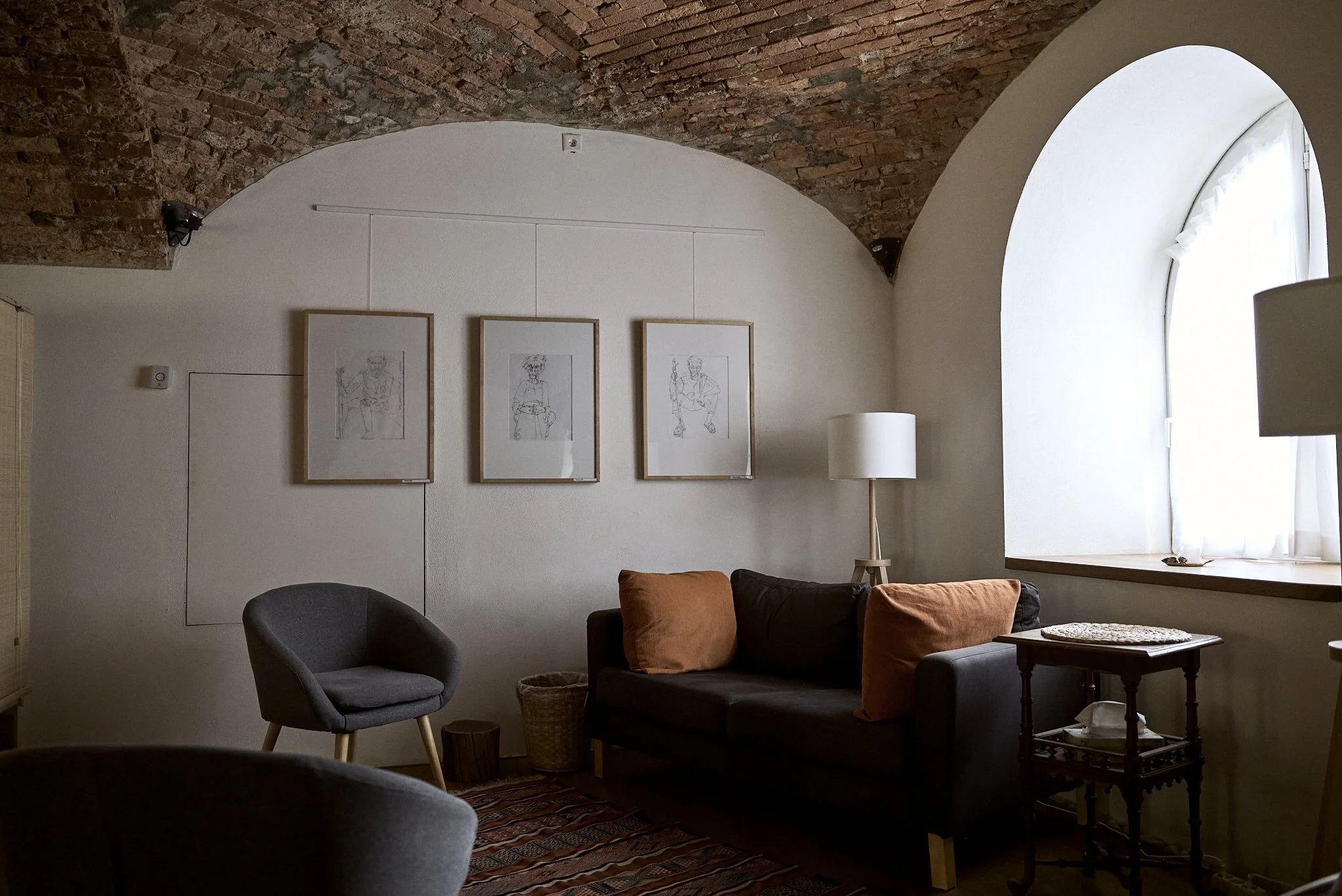 Ein gemütliches Wohnzimmer mit dunklem Sofa, einem grauen Sessel, einem runden Teppich, einer Stehlampe, Kunstwerken an der Wand und einem großen Fenster mit weißen Vorhängen.