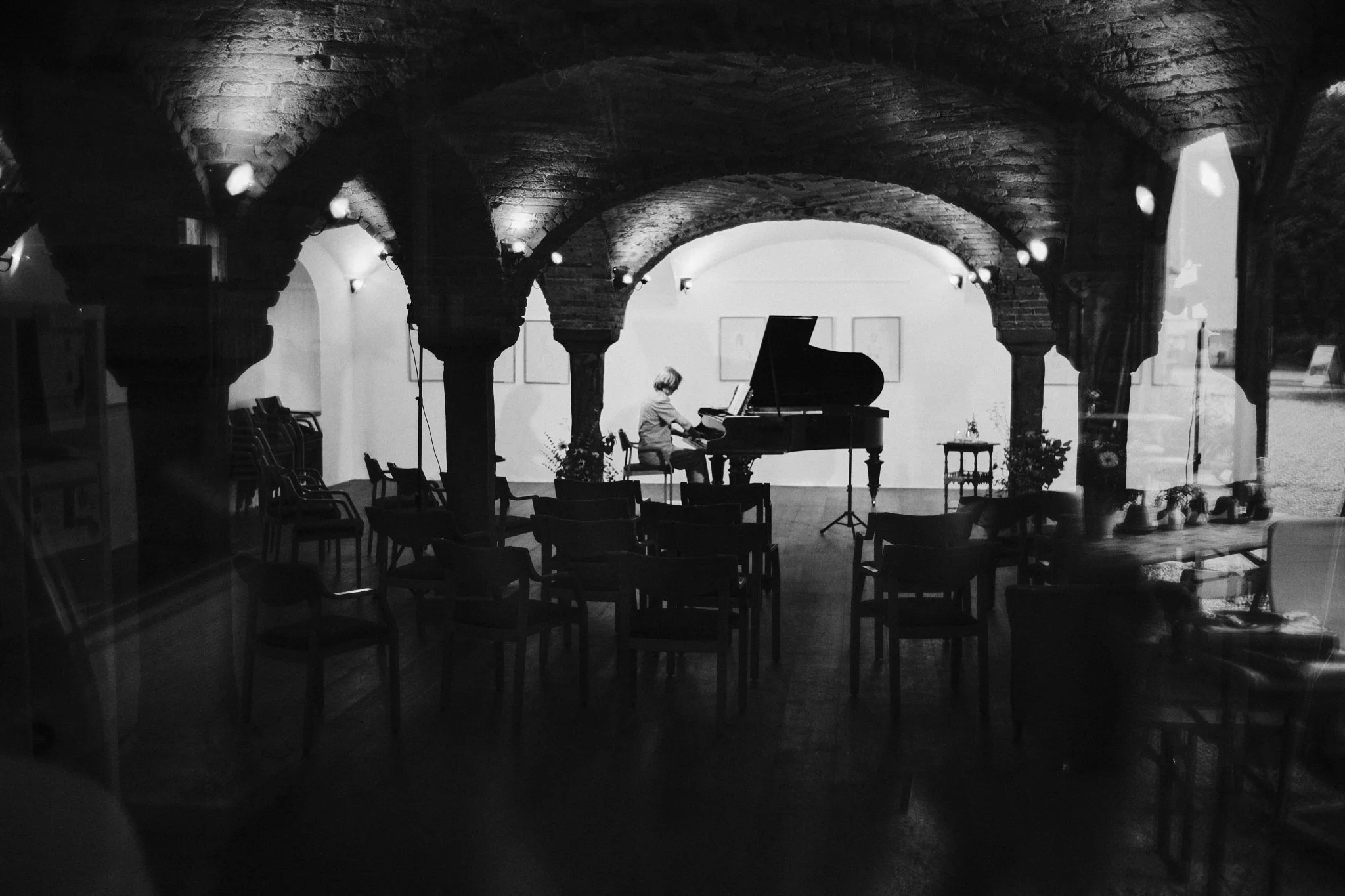 Junge Person spielt Klavier in einem dunklen Raum mit gewölbtem Brickdecke, Sitzgelegenheiten und Pflanzen, durch ein Fenster sichtbar.