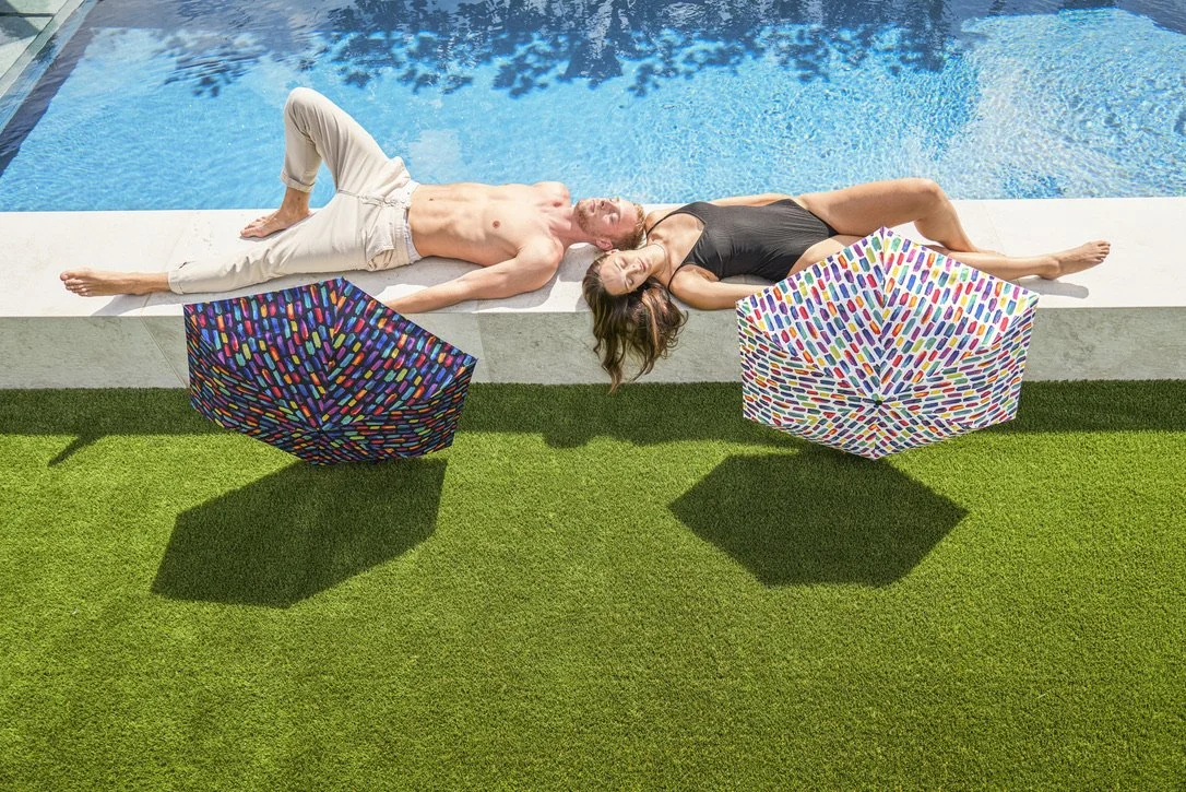 Ein Mann und eine Frau liegen entspannt nebeneinander auf der Poolkante, während bunte Regenschirme Schatten auf den Rasen werfen.