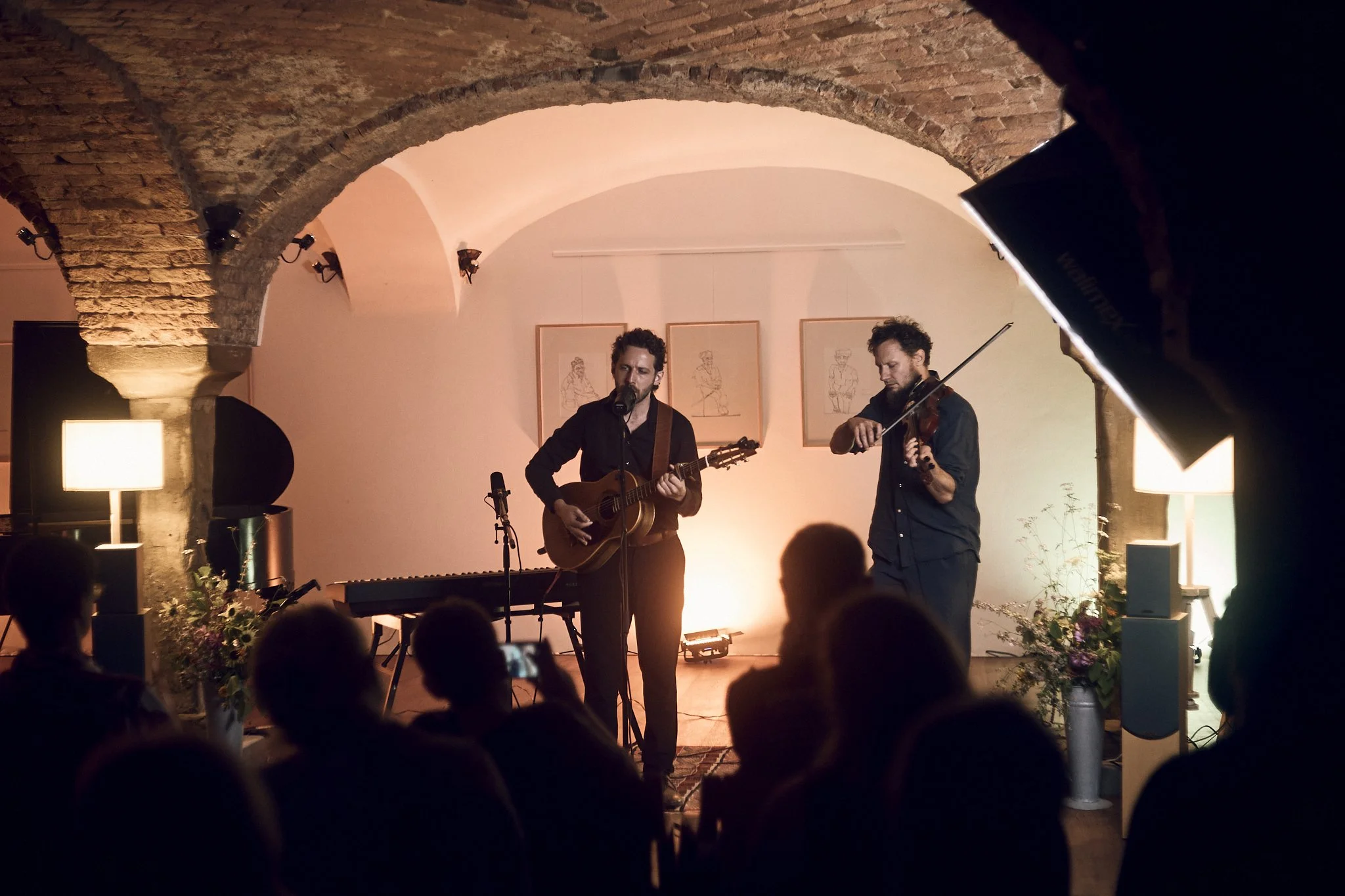 Zwei Musiker spielen im Kulturraum Oberhofen mit Steinbögen, einer mit Gitarre und der andere mit Violine, vor einem Publikum.