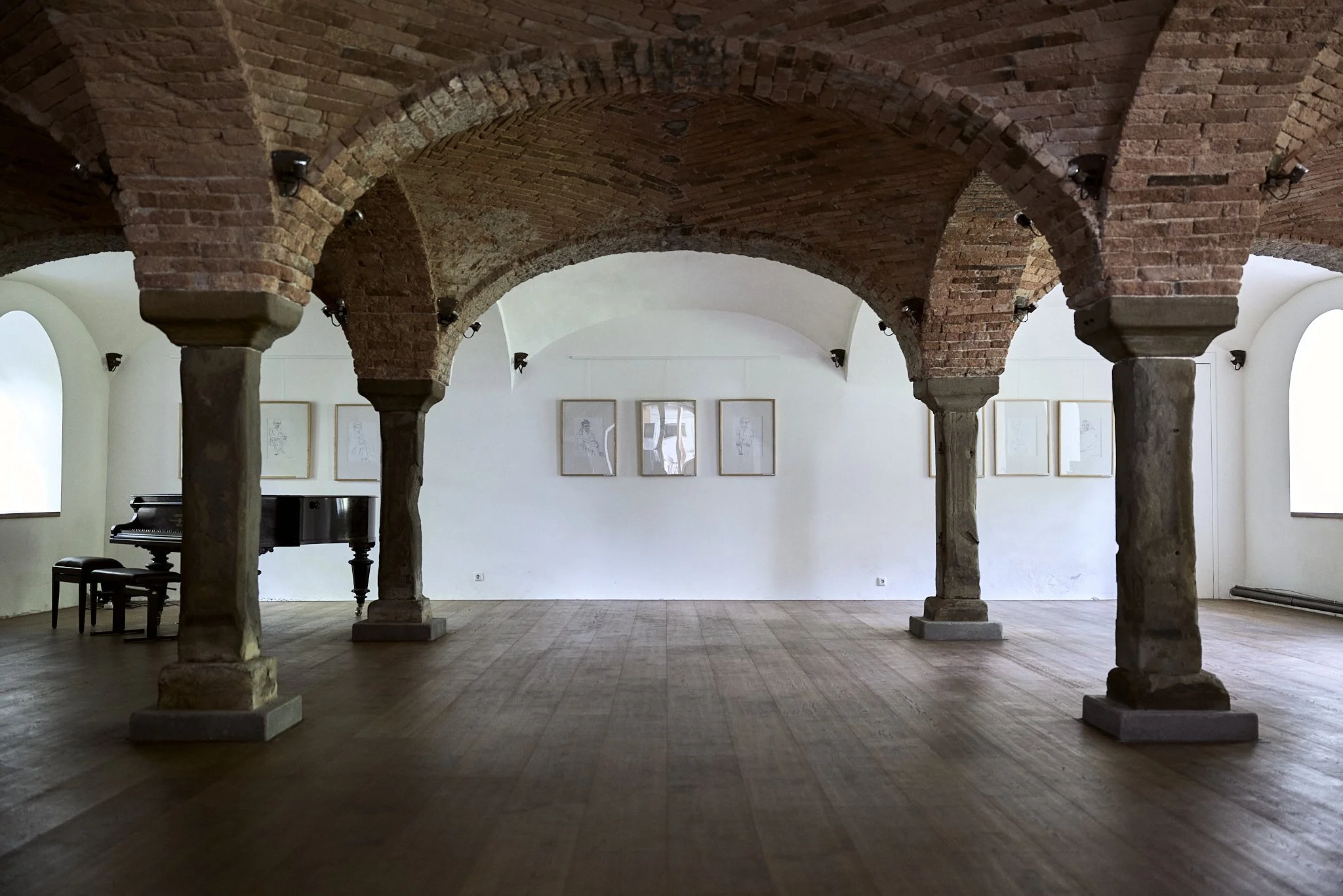 Ein leerer Raum mit Holzboden, weißen Wänden, großen Fenstern, einer schwarzen Konzertflöte und Bildern an der Wand, mit einer sichtbaren brick arched Decke. Kulturraum Oberhofen Ausstellung