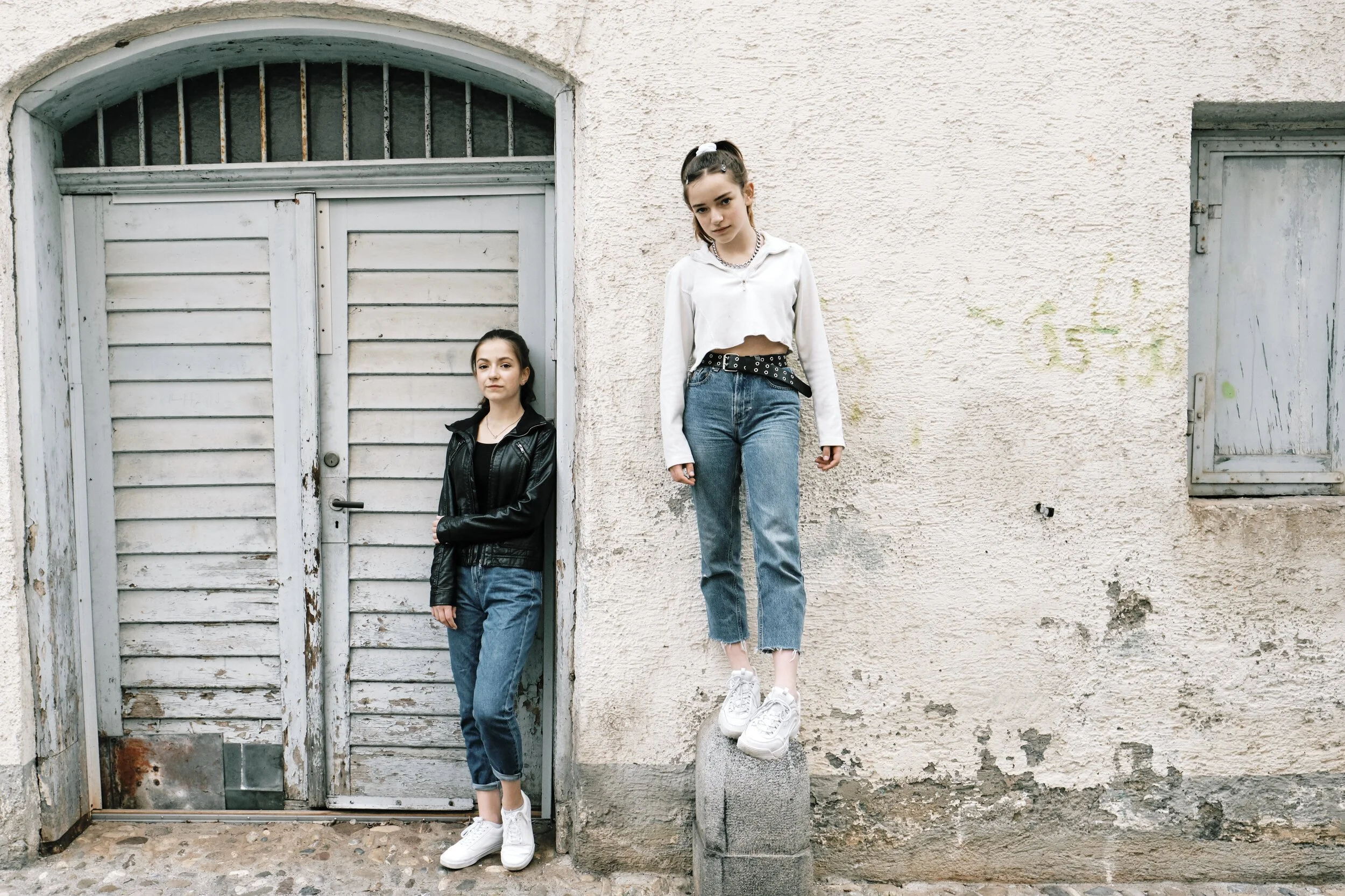 Zwei junge Frauen stehen an einer alten, weißen Wand in einer Straße, eine lehnt an einer Tür und die andere steht auf einem Stein, beide tragen Jeans und weiße Turnschuhe.