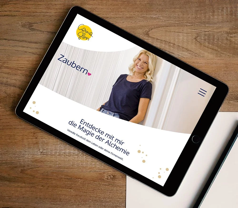 Tablet auf Holz mit Website, die eine blonde Frau lächelnd zeigt, Überschrift 'Entdecke mit mir die Magie der Alchemie', Logo in Gelb, und der Titel 'Zaubern ♥'