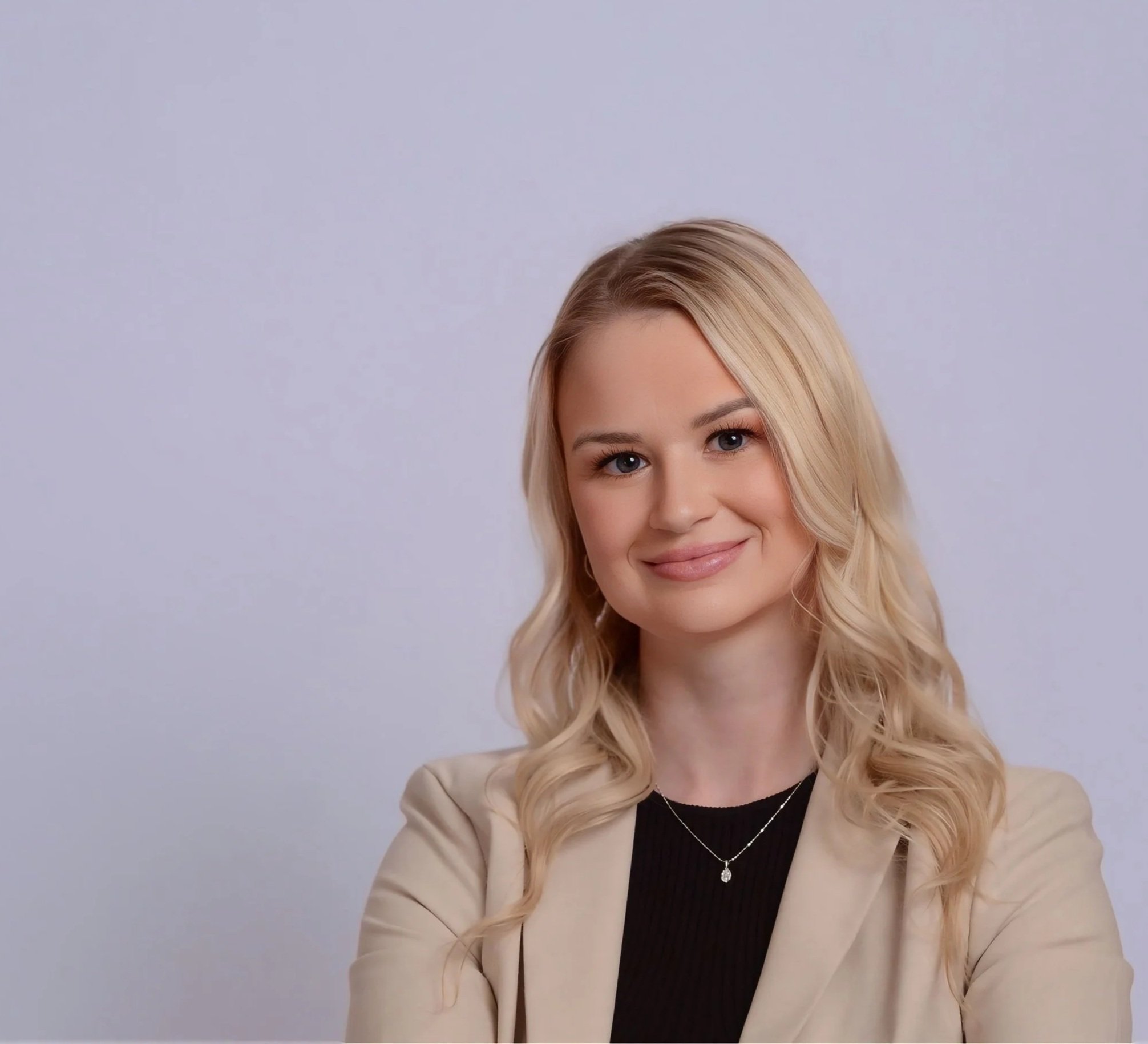 Portrait of a young woman with blonde, wavy hair, smiling slightly, dressed in a beige blazer and black top, standing against a plain, light-colored background.