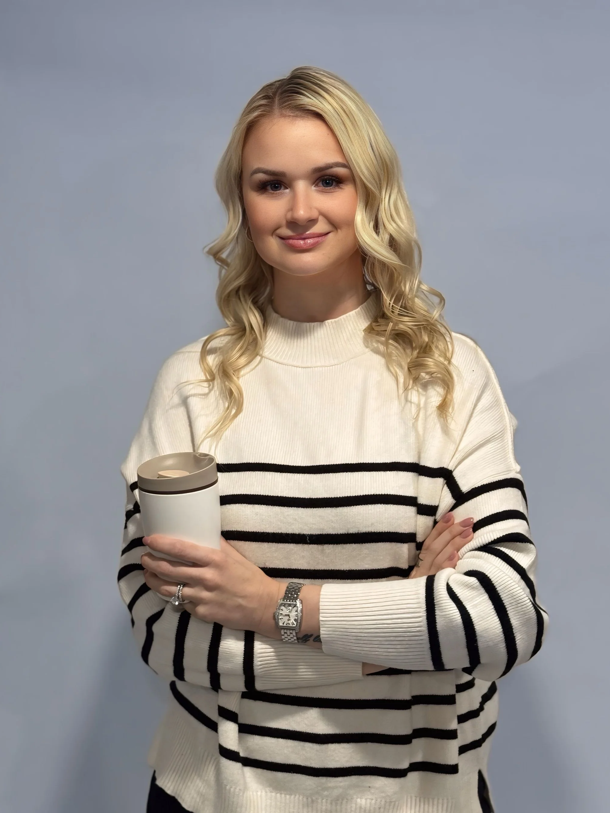 A woman with long blonde hair, wearing a cream-colored sweater with black stripes, is smiling and holding a travel mug. She is standing against a plain gray background.