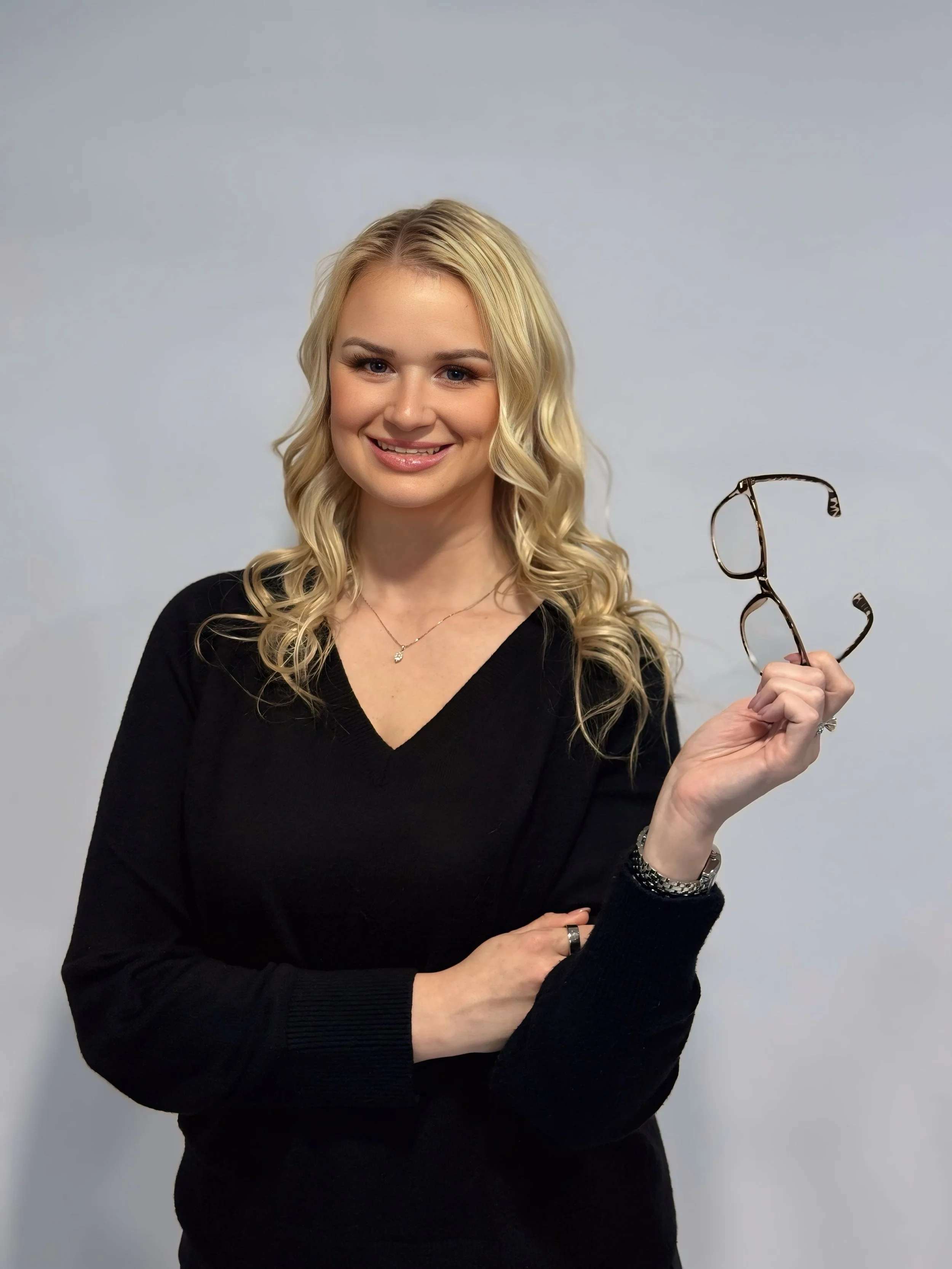 A young woman with blonde curly hair wearing a black sweater holding a pair of glasses and smiling at the camera against a plain light gray background.