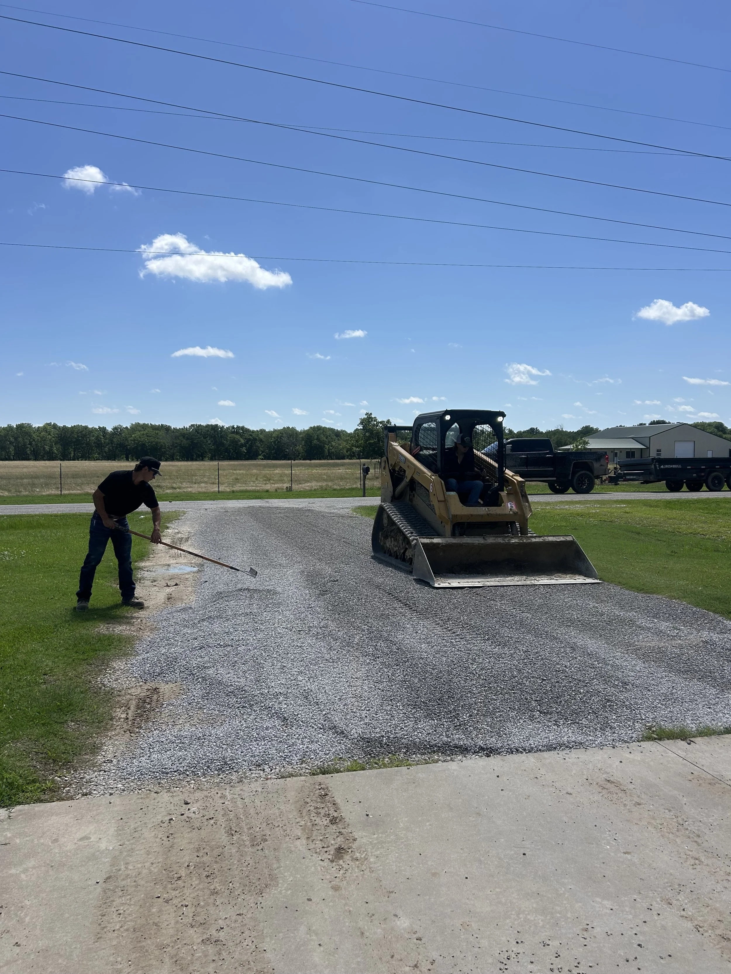 Two men are working on a gravel driveway; one man is raking gravel while the other man is operating a small bulldozer. The scene is outdoors on a sunny day with blue skies and a few clouds.