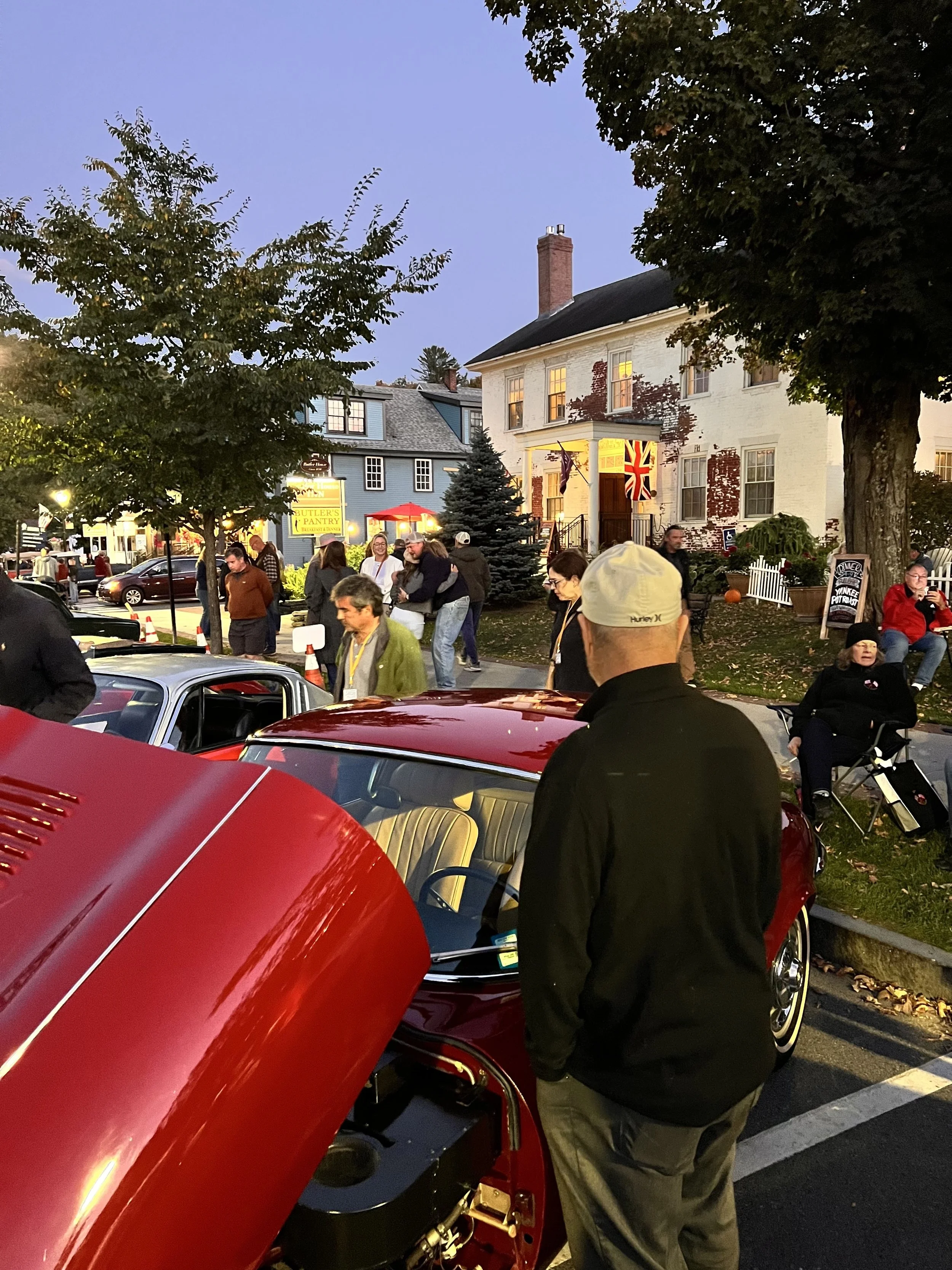 People gathered around a red vintage car at an outdoor event during the evening, with a house and trees illuminated by warm lights in the background.