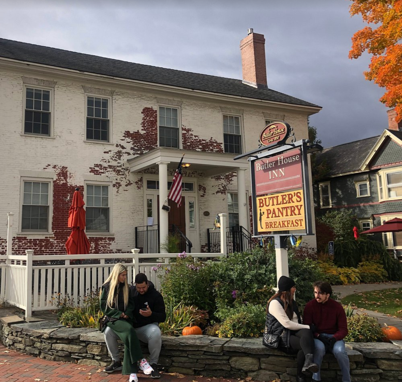 People sitting on a stone wall outside the Butler House Inn and Butler's Pantry Breakfast restaurant, a white brick building with peeling paint and a porch. There are pumpkins and fall decorations, and a sign displaying the restaurant name.