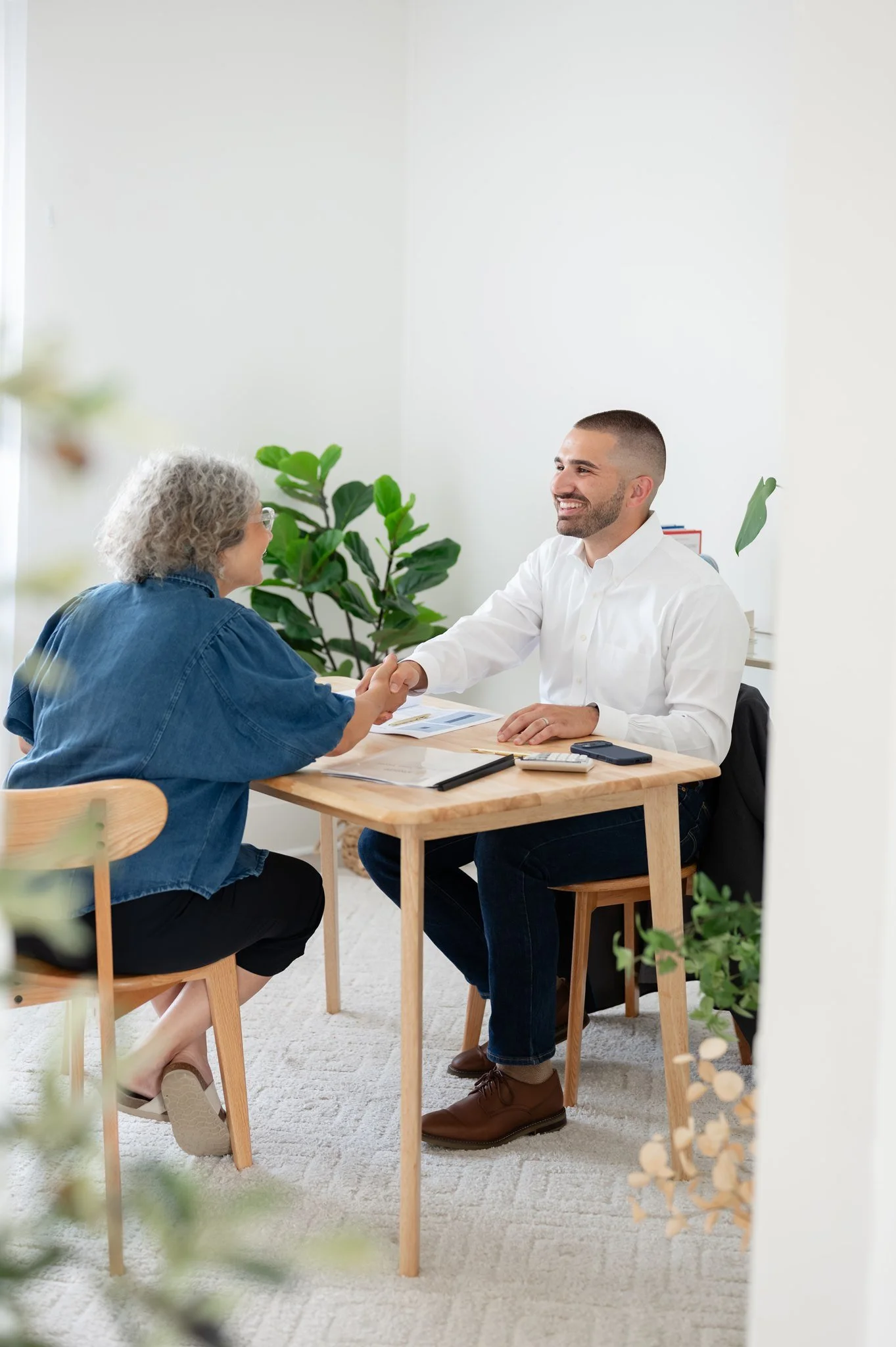 A young man in a white shirt and brown shoes shaking hands with an older woman in a blue denim jacket, sitting at a wooden table in a bright room with green plants.