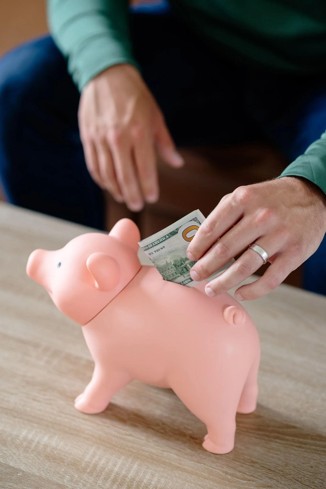 Person inserting money into a pink piggy bank on a wooden surface.