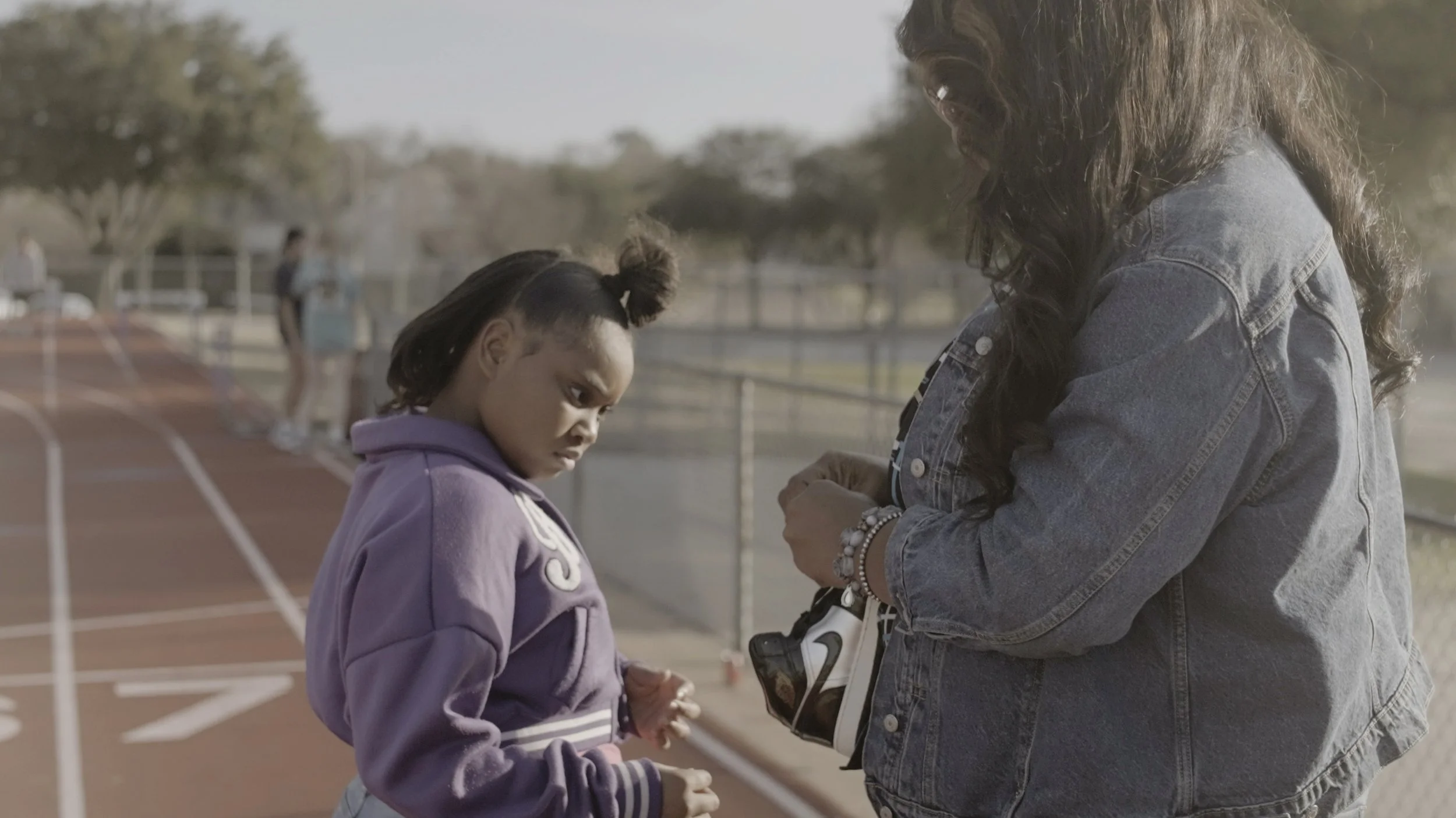 A girl and a woman standing near a running track, with the girl looking serious and the woman holding a pair of shoes. In the background, other people are seen, some talking and walking.