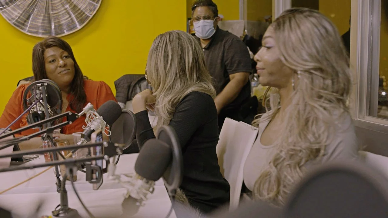 Four women seated at a table with microphones, engaged in conversation. One woman is wearing an orange top, another has blonde hair and is wearing a black blazer, the third woman has long blonde hair and is wearing a white top, and the fourth woman in the background is wearing a mask and dark clothing. The room has yellow walls and a large clock on the wall.