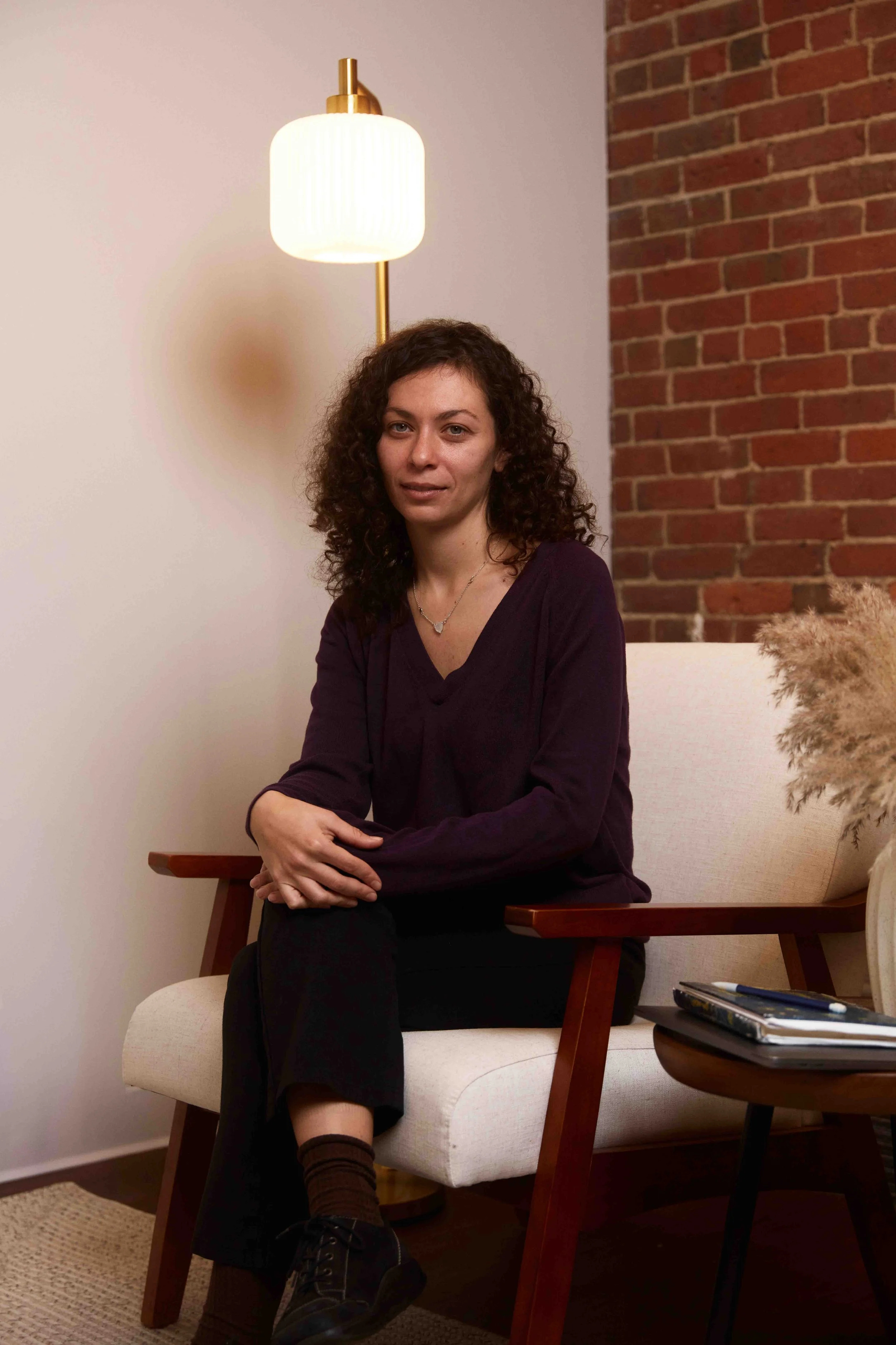 A woman with curly brown hair sitting on a wooden frame cream-colored armchair, wearing a dark purple top, black pants, socks, and shoes, in a room with a brick wall, white wall, a standing lamp, and a side table with a notebook and pens.