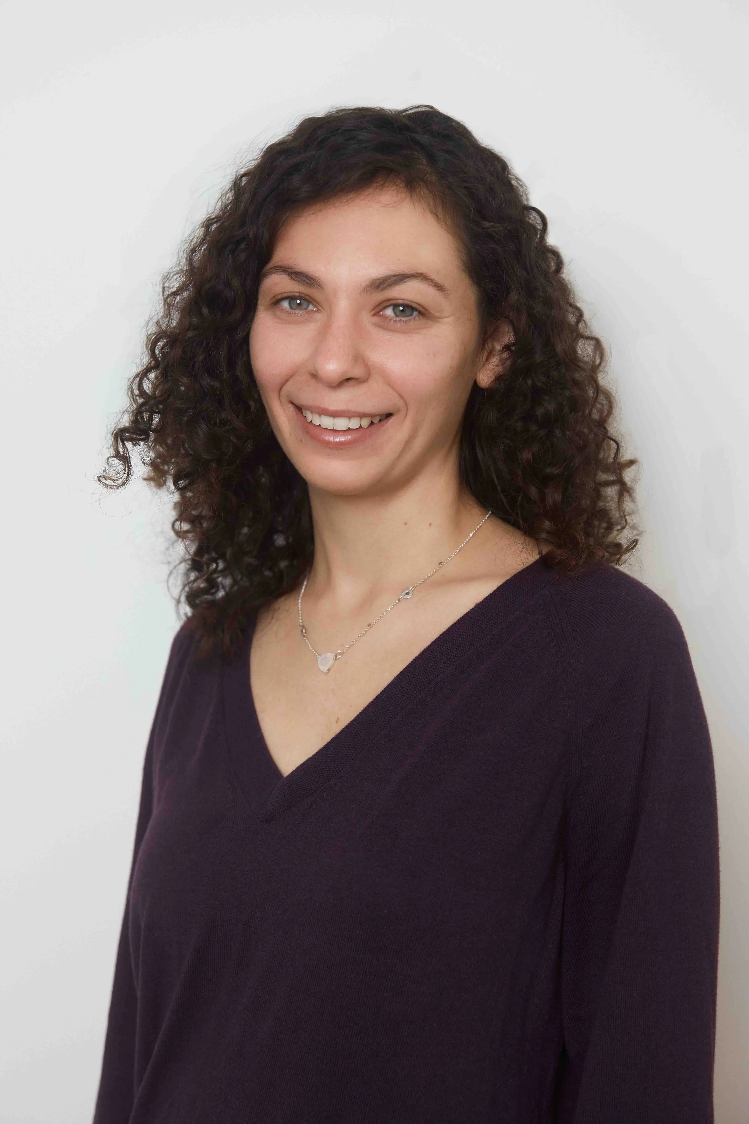 A woman with curly brown hair wearing a purple V-neck sweater and a silver necklace, smiling against a plain white wall.