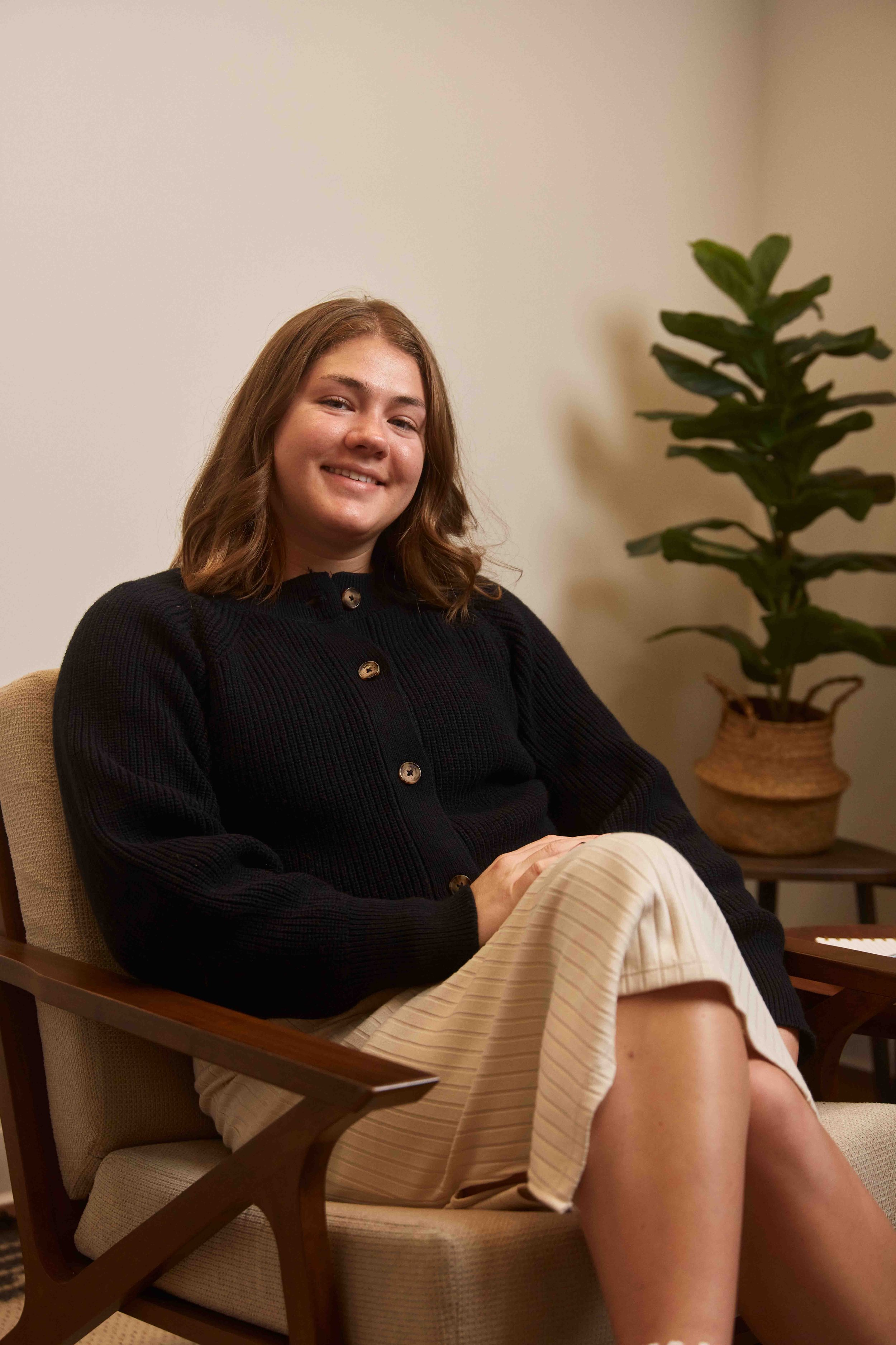 A young woman with shoulder-length brown hair smiling and sitting on a beige armchair in a room with a potted plant in the background.