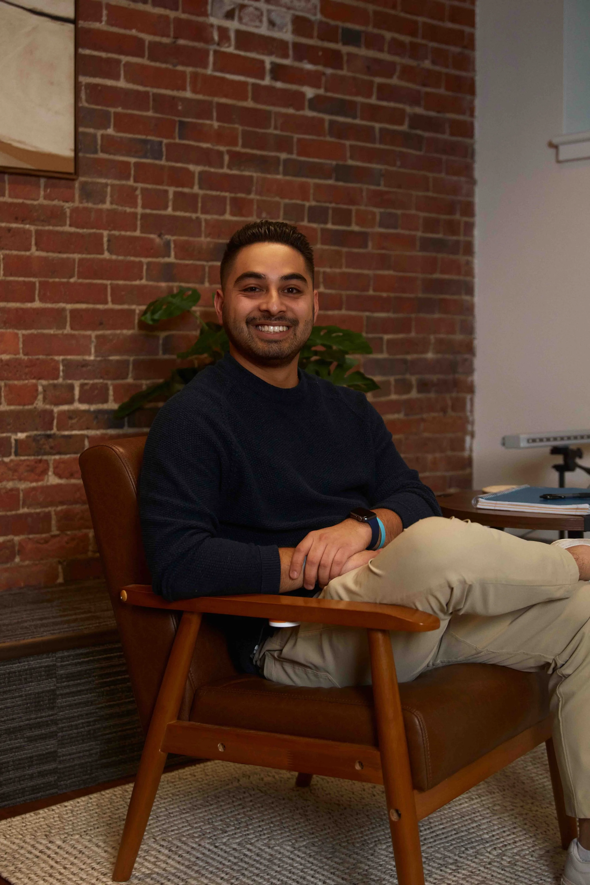 A smiling man sitting in a brown leather armchair in a room with a brick wall, a plant behind him, and a table with books and a whiteboard in the background.