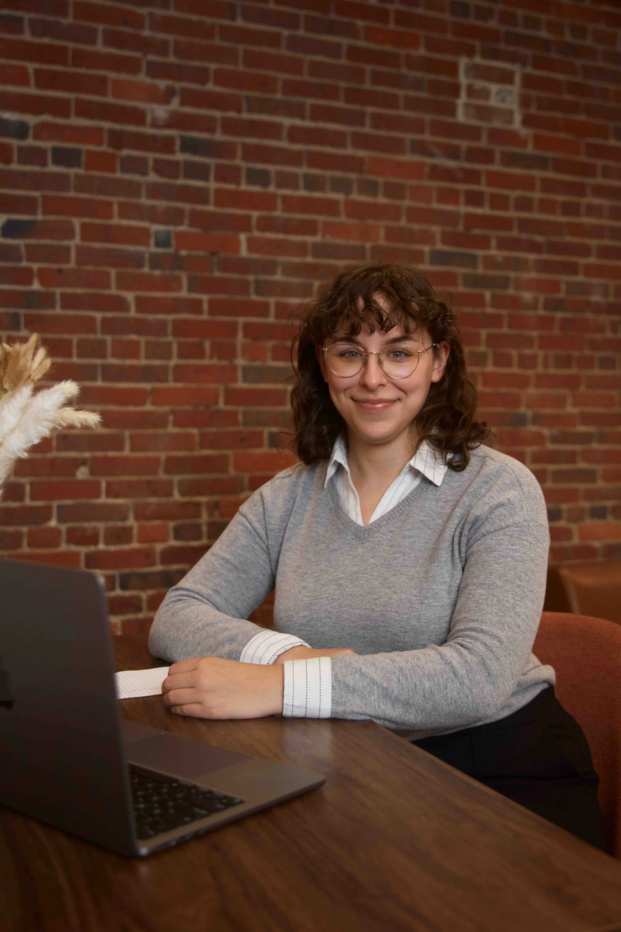 A woman with curly brown hair and glasses sitting at a wooden table with a laptop, in front of a brick wall, smiling at the camera.