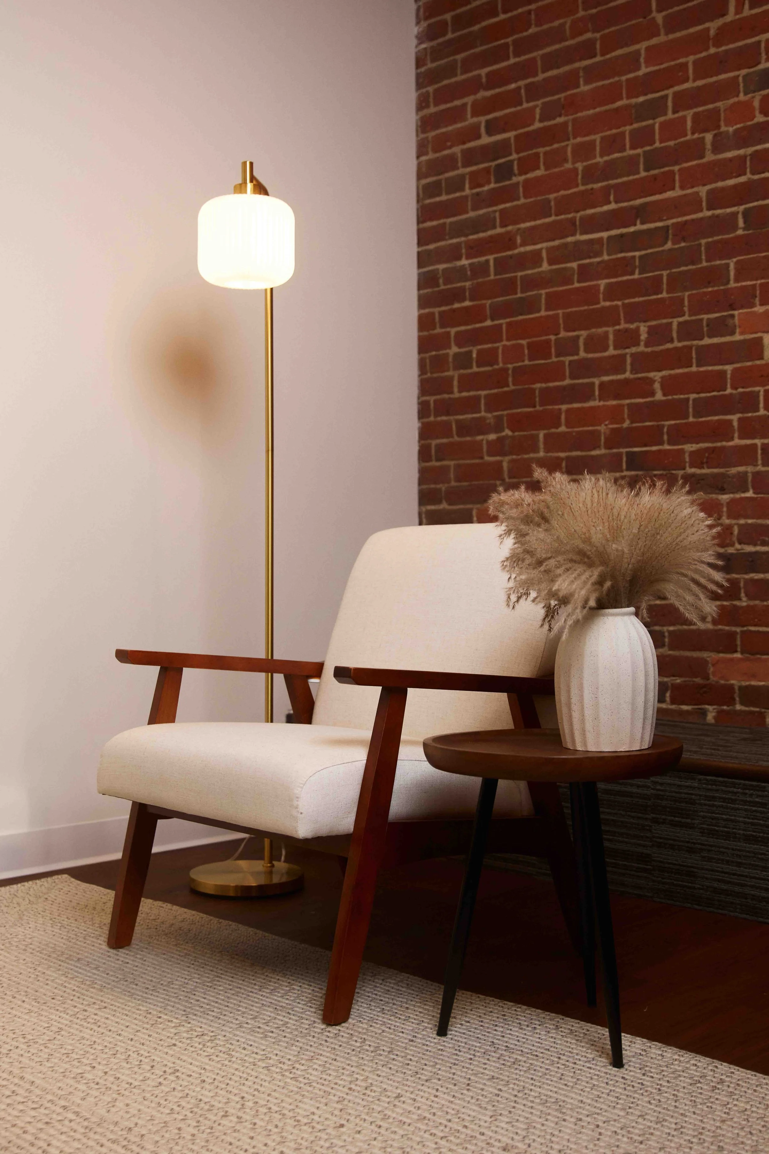 A living room corner with a white upholstered armchair featuring wooden armrests, a round wooden side table with a white vase filled with dried pampas grass, a tall floor lamp with a white shade and brass stand, a brick accent wall, and a beige rug on a wooden floor.