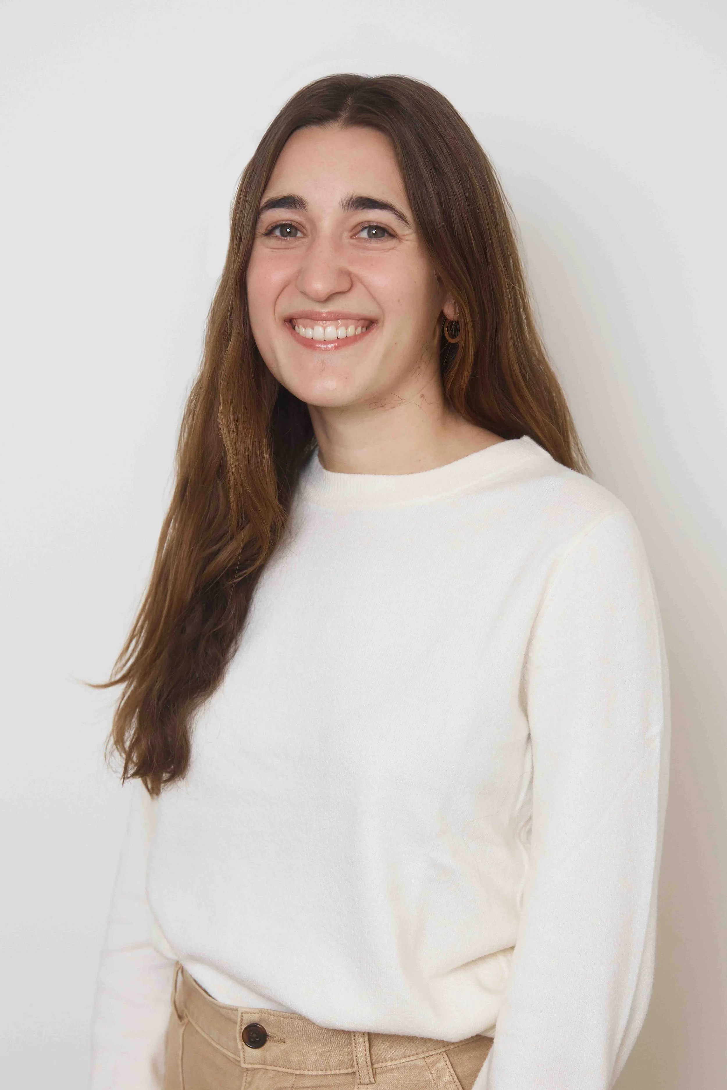 A young woman with long brown hair, wearing a white sweater and beige pants, smiling while standing against a plain white background.